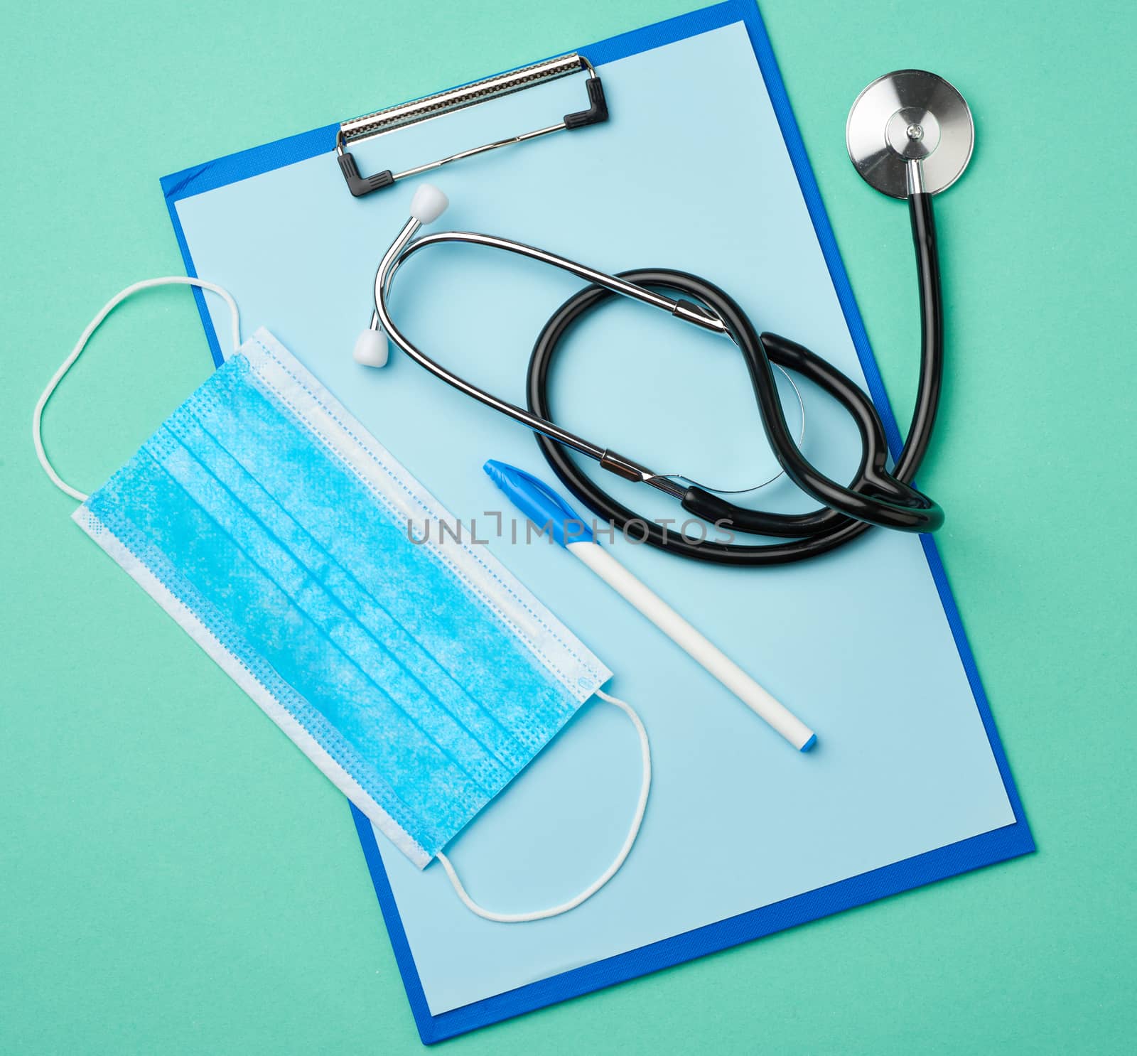 metal stethoscope, paper holder with a blank blue sheet, disposable blue medical mask on a green background, top view, flat lay