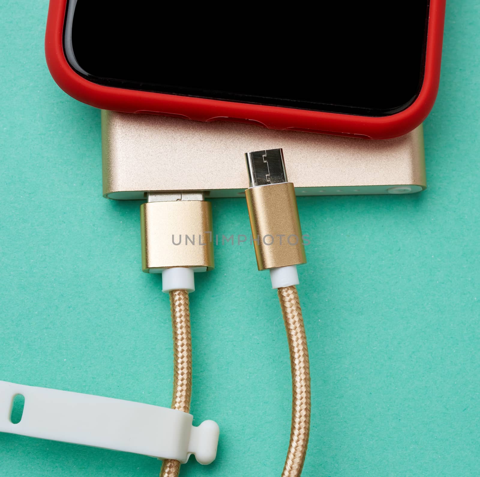red smartphone with a blank black screen is connected by a cord to a power bank, green background, top view