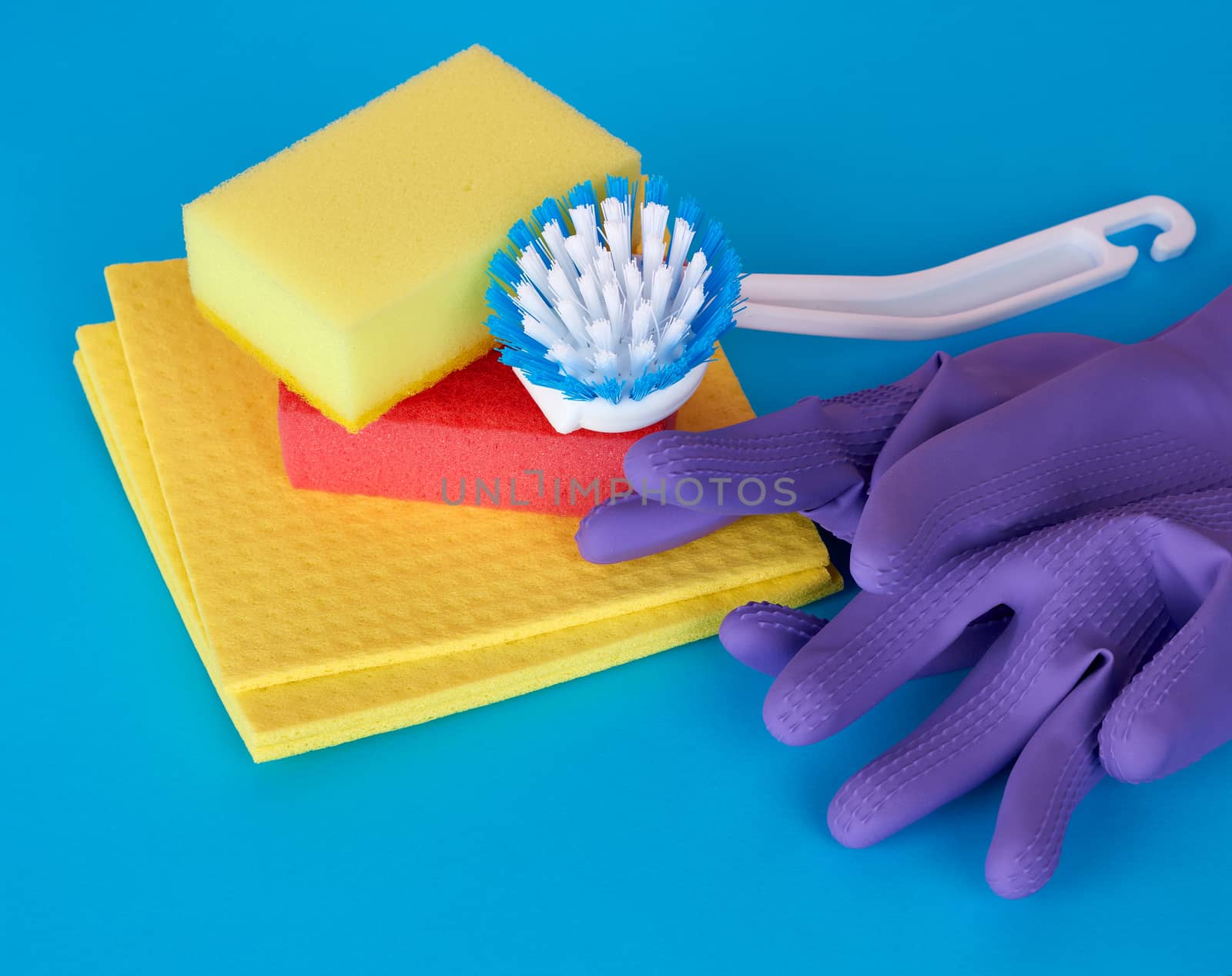 multi-colored kitchen sponge, white plastic brush, rubber purple gloves on a blue background, set of items for cleaning