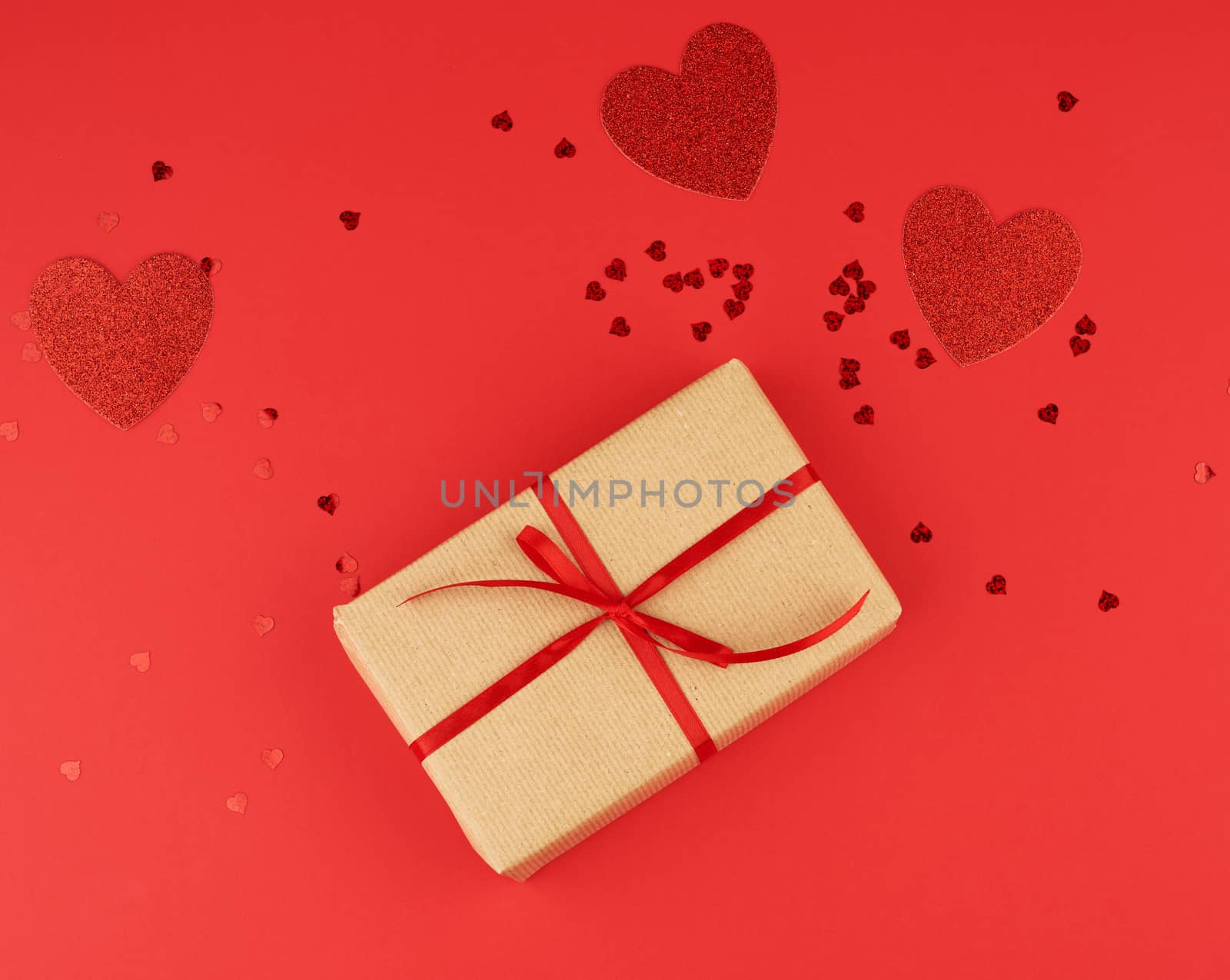box wrapped in brown kraft paper and tied with a red thin silk ribbon, scattered confetti in the shape of hearts, red background, festive backdrop for Valentine's Day