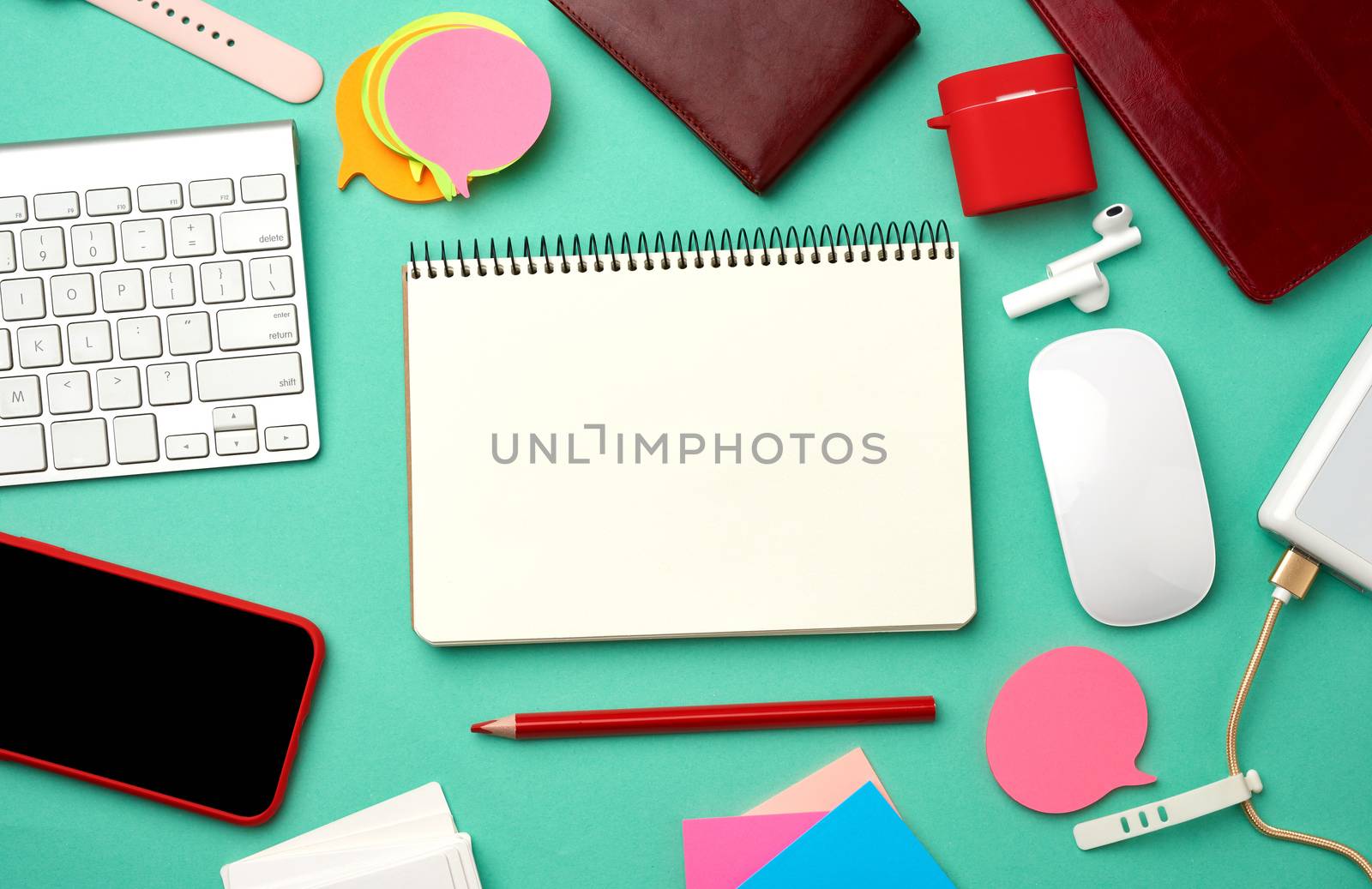paper notebook, keyboard, power bank with cable, red smartphone with blank black empty screen, headphones, blank paper business cards and wireless mouse on green background, workplace, top view