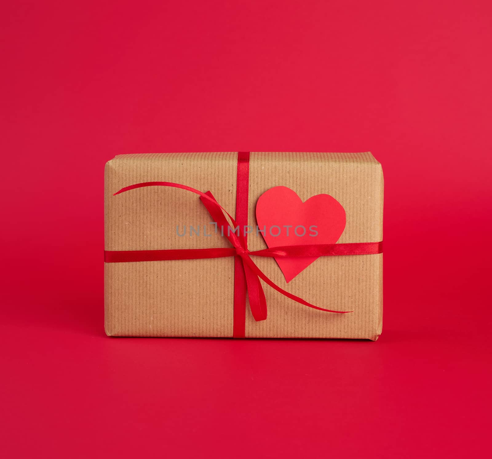 box wrapped in brown kraft paper and tied with a red thin silk ribbon, cut out of paper hearts, red background, gift for Valentine's Day, birthday, flat lay