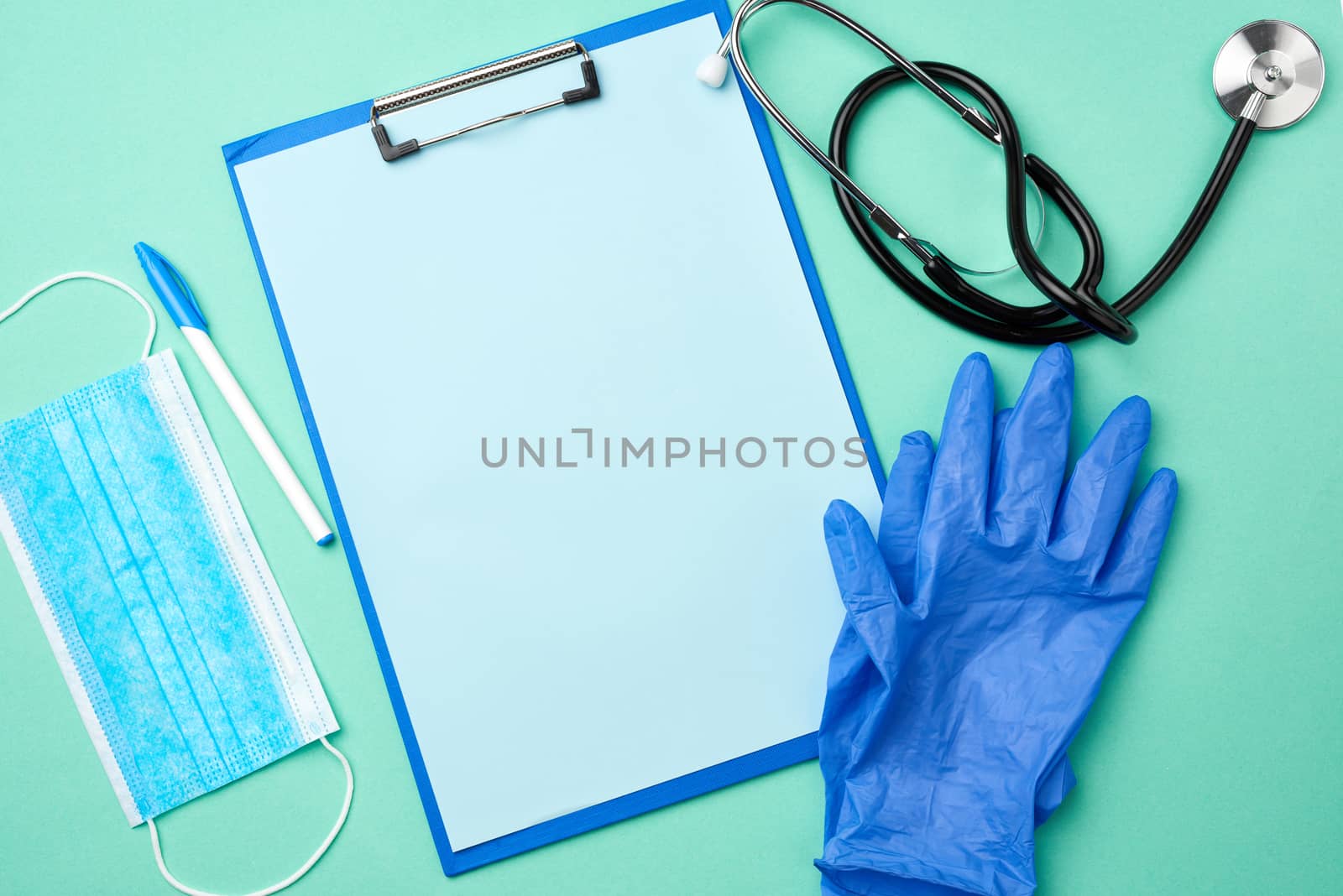 metal stethoscope, paper holder with a blank blue sheet, disposable blue medical mask on a green background, top view, flat lay