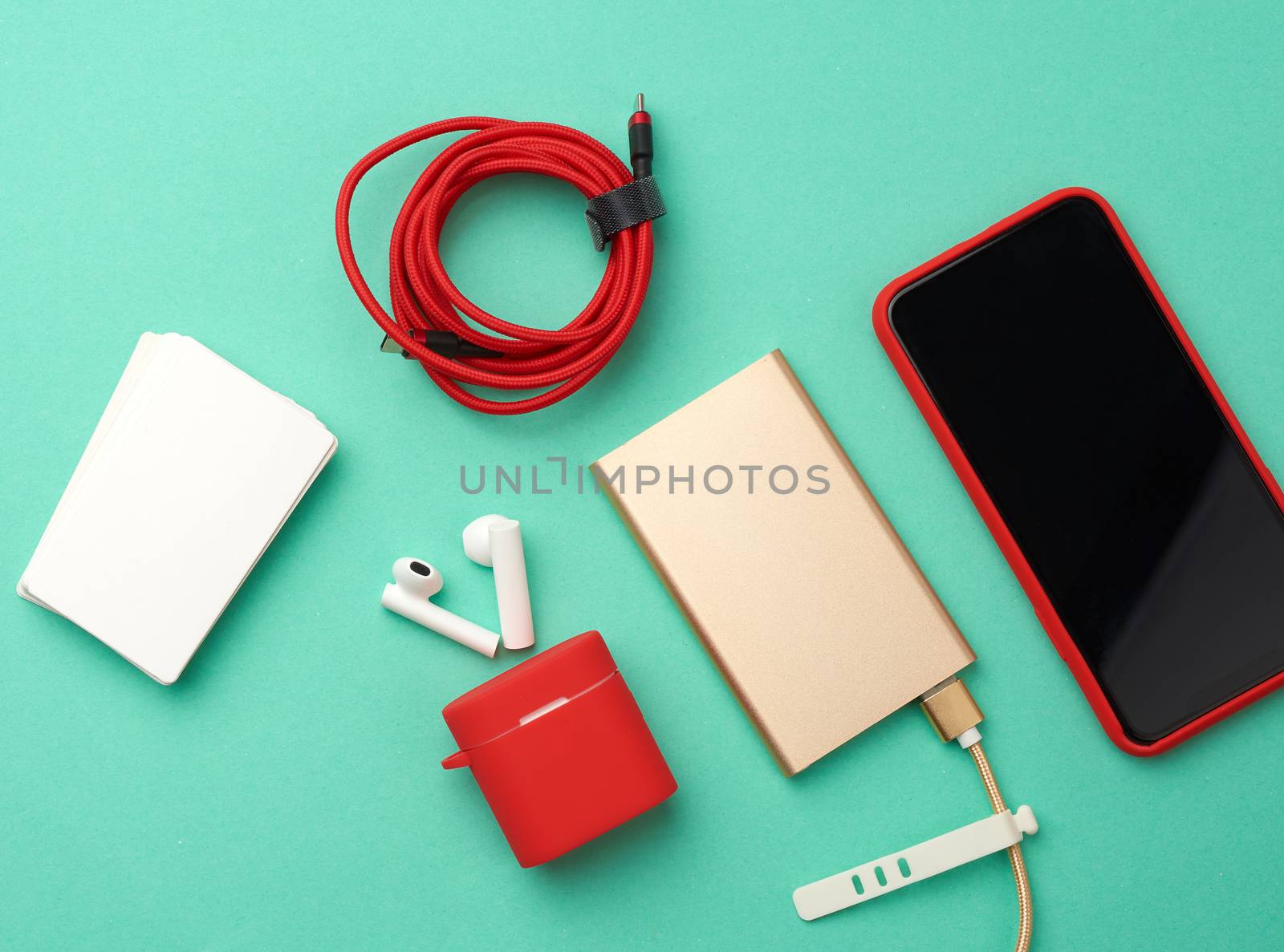 golden power bank with cable, red smartphone with blank black screen, box with headphones, empty paper business cards on green background, top view
