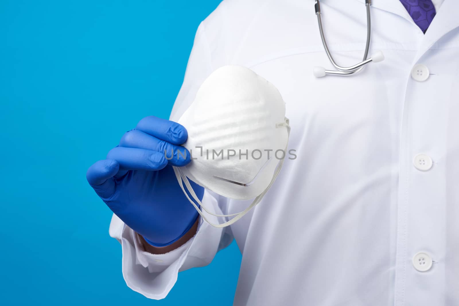 male doctor in blue latex sterile gloves holds a white disposable mask made of non-woven material on a blue background, protective accessory for the respiratory tract from the virus
