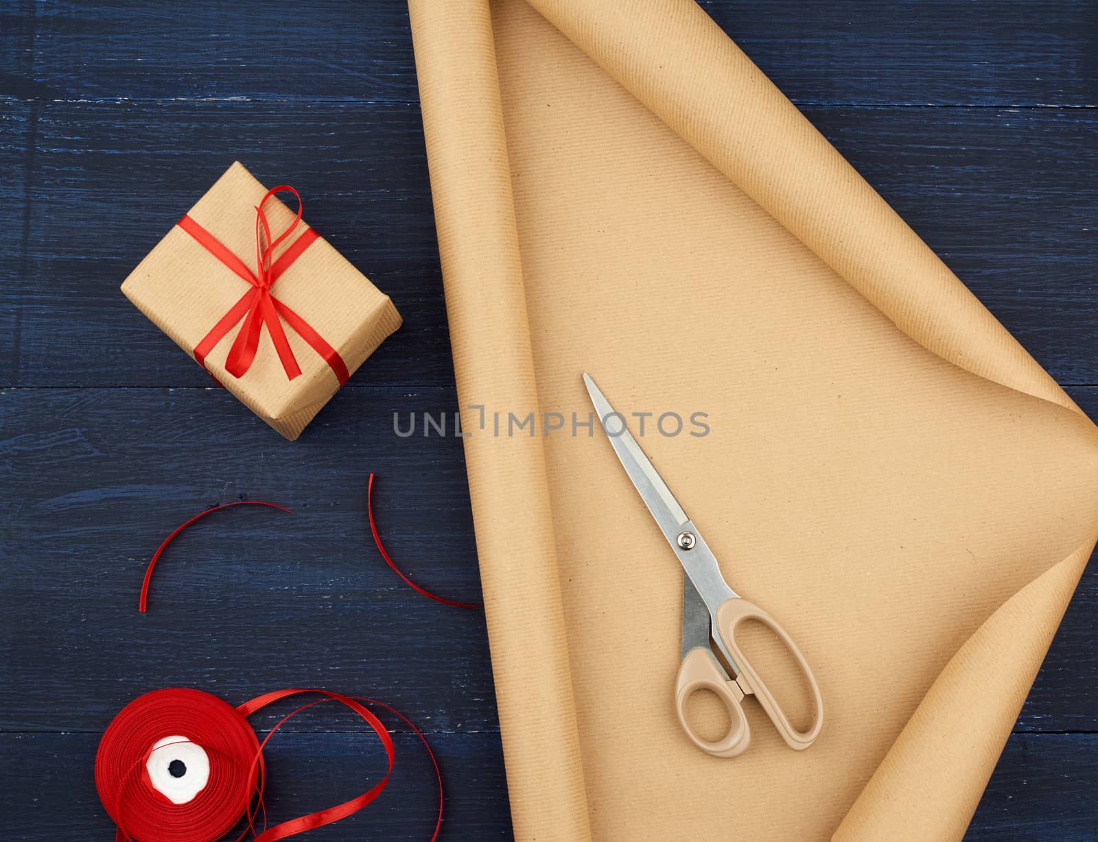 roll of brown kraft packaging paper, box tied with a red silk ribbon, scissors and a bobbin with ribbon on a blue wooden background, top view
