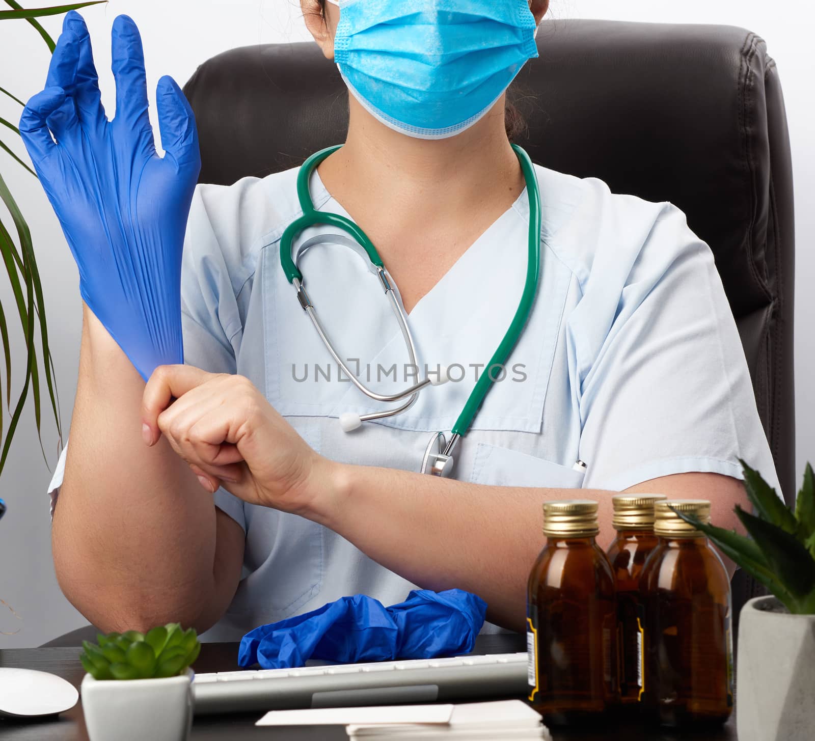 woman doctor in a blue uniform, disposable medical mask is sitting in a chair at the table and puts on his hands blue sterile latex gloves, doctor’s office