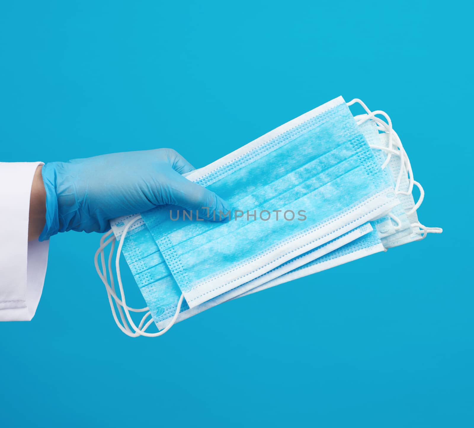doctor in a white coat, blue latex sterile gloves holds textile disposable medical masks in his hand on a blue background, protective accessory for the respiratory tract from the virus