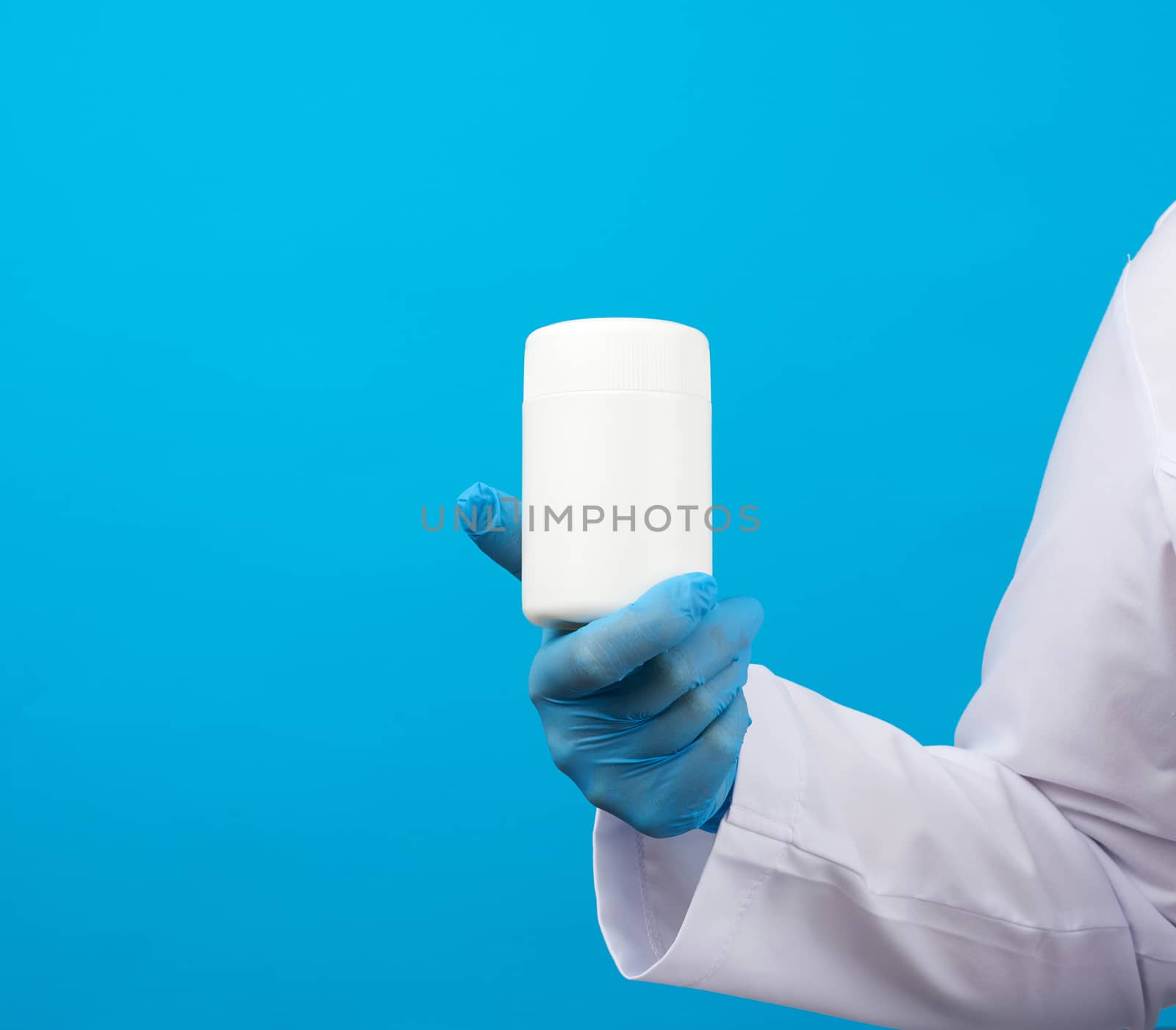 hand in blue sterile gloves holds a white plastic jar for pills, medical treatment concept for diseases, blue background