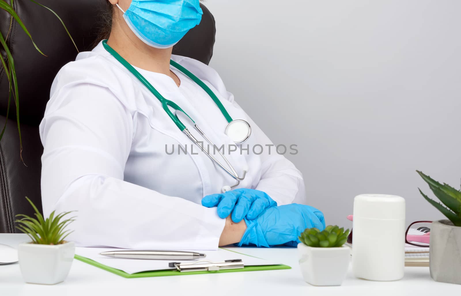 doctor in a white coat and blue latex gloves sits at a white work table in his office, concept of patient reception in the clinic