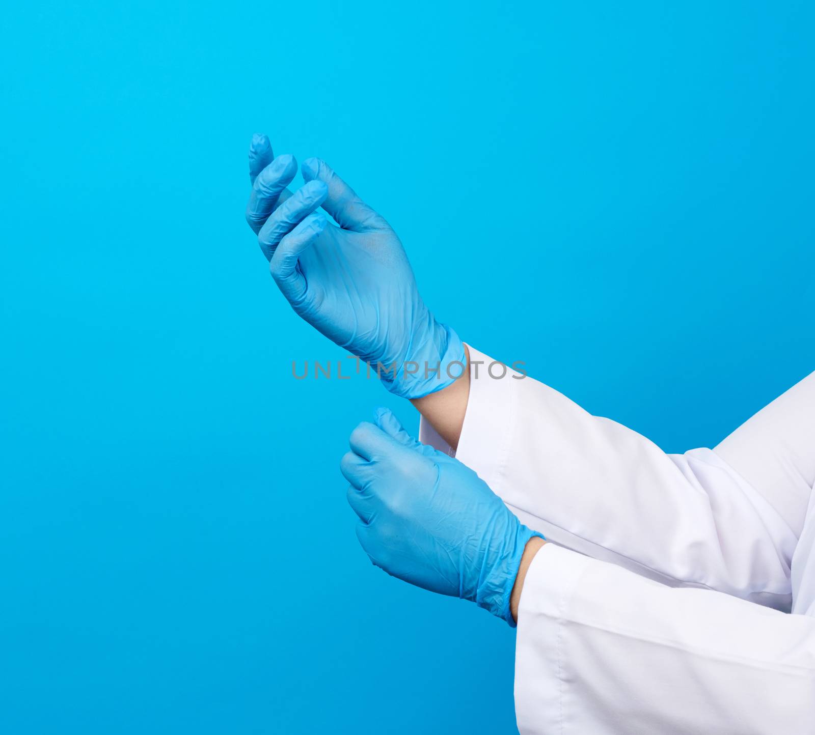 female doctor in a white coat puts on her hands blue latex sterile medical gloves on her hands, blue background, close up