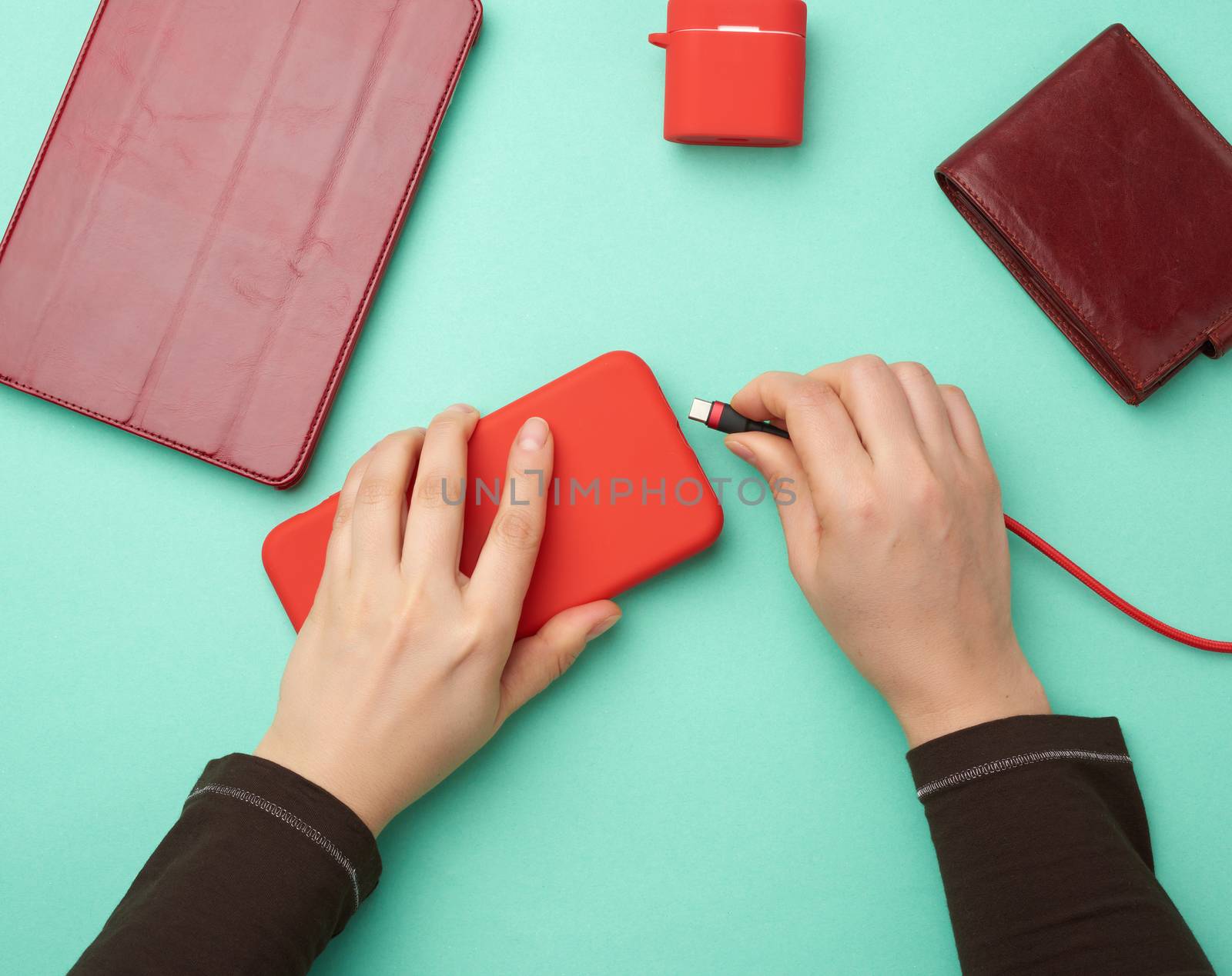 two hands holding a red mobile smartphone and a charging cable on a green background, top view
