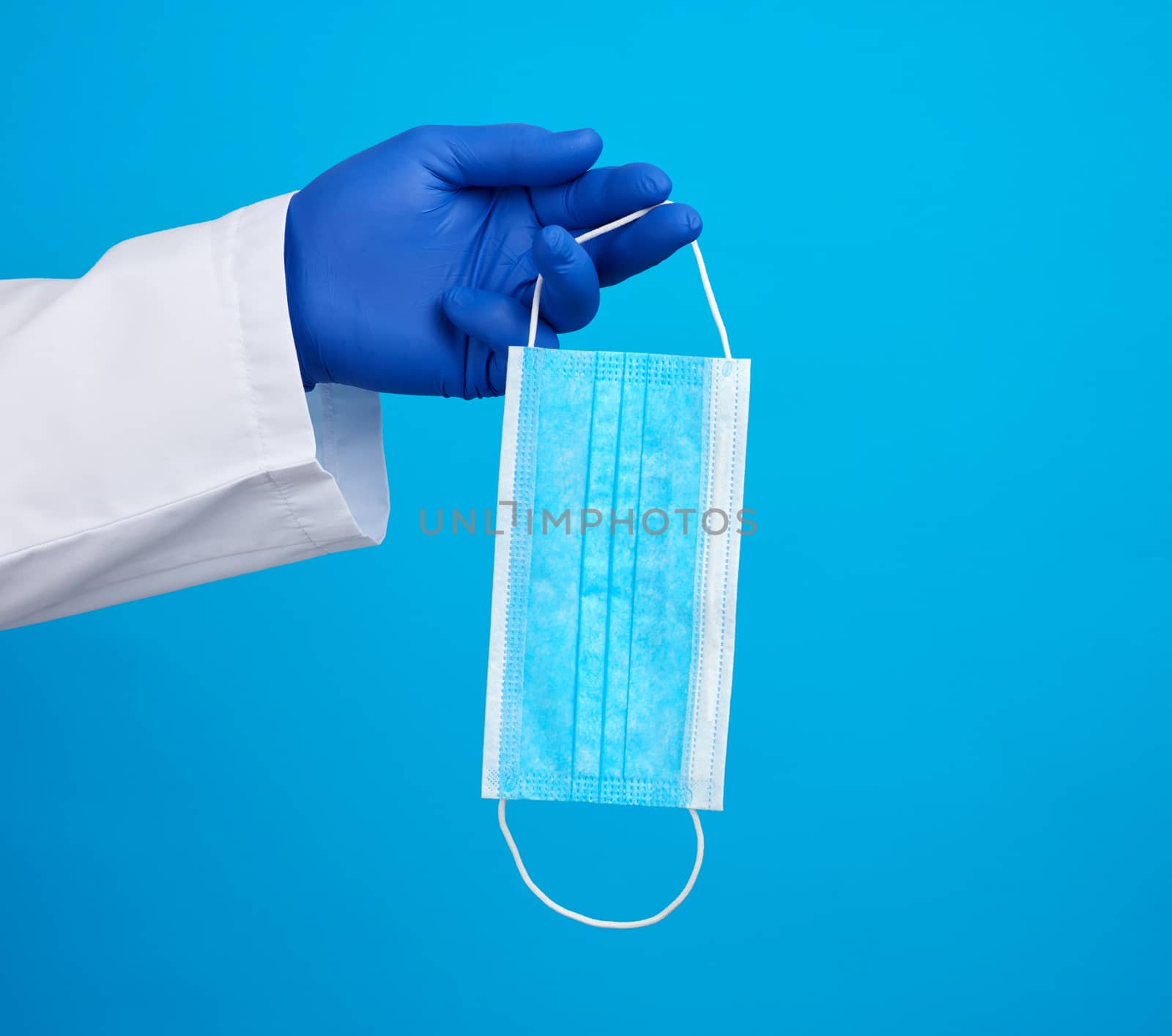 doctor in a white coat, blue latex sterile gloves holds textile disposable medical masks in his hand on a blue background, additional protection against viruses during an epidemic, pandemic