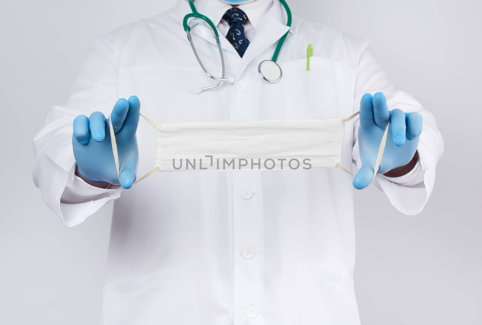 doctor in a white coat, blue latex sterile gloves holds textile medical masks in his hand on a white background, homemade mask made of white gauze and elastic
