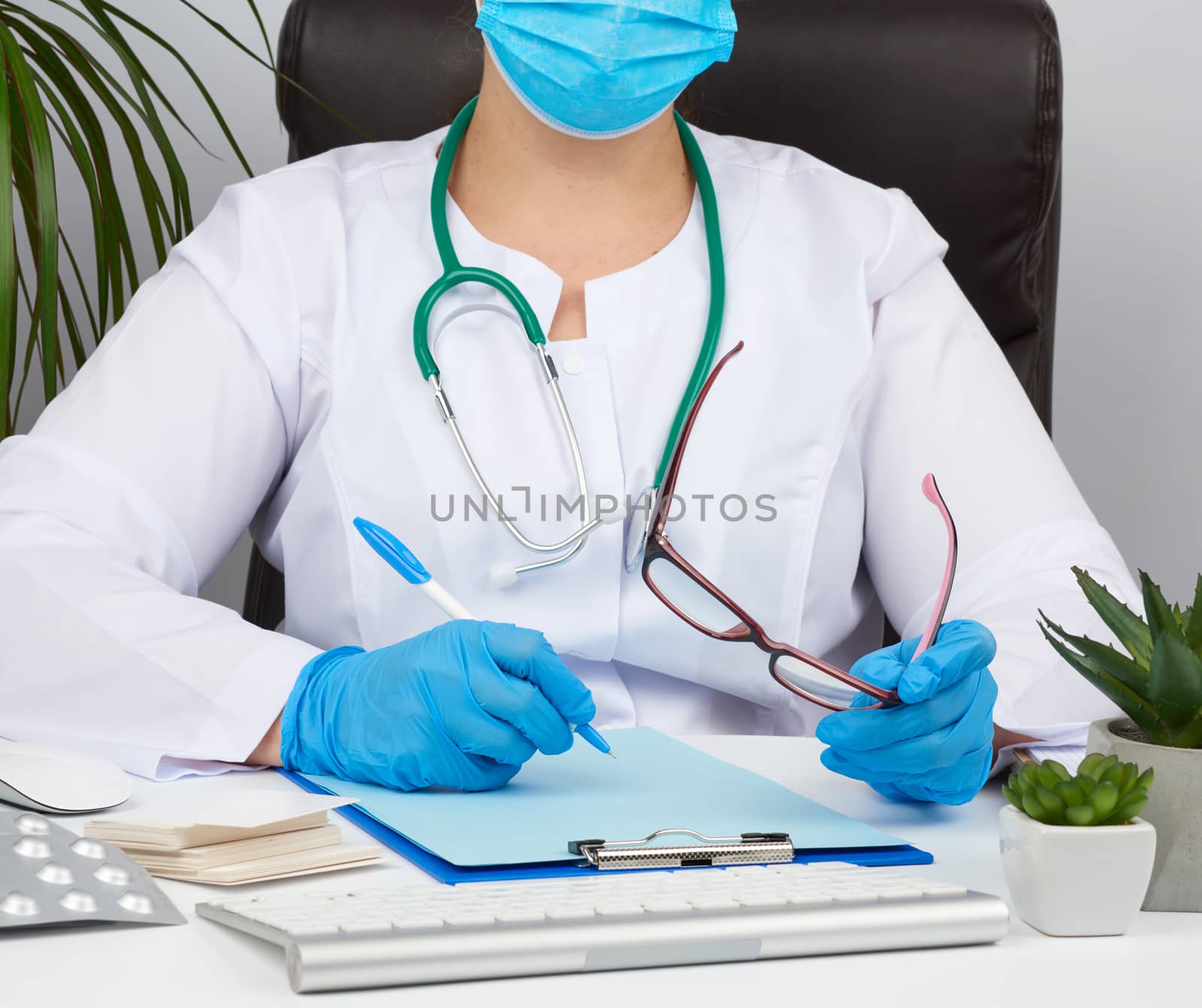 young woman doctor in a white coat, sterile medical gloves writes a prescription on a form, therapist sits on a chair at a white table, workplace