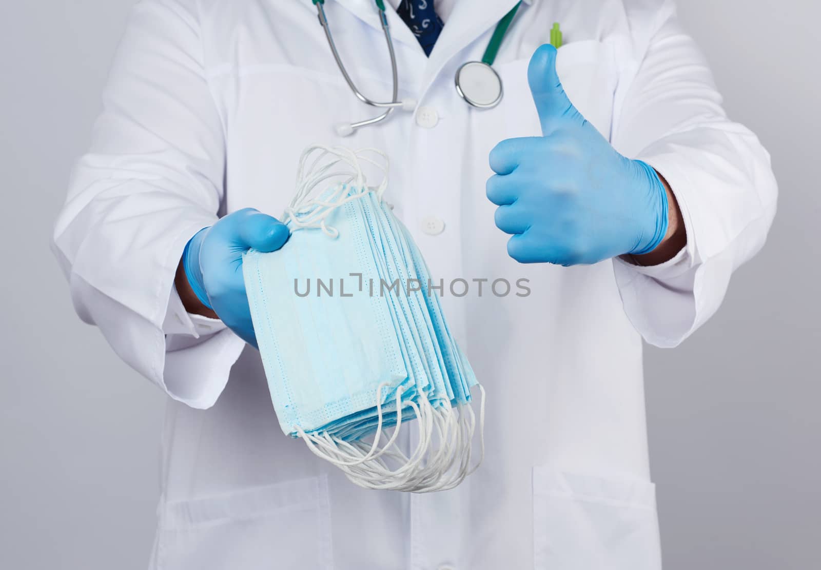 doctor in a white coat, blue latex sterile gloves holds textile medical masks in his hand on a white background, other hand shows like, protective accessory against viruses and bacteria
