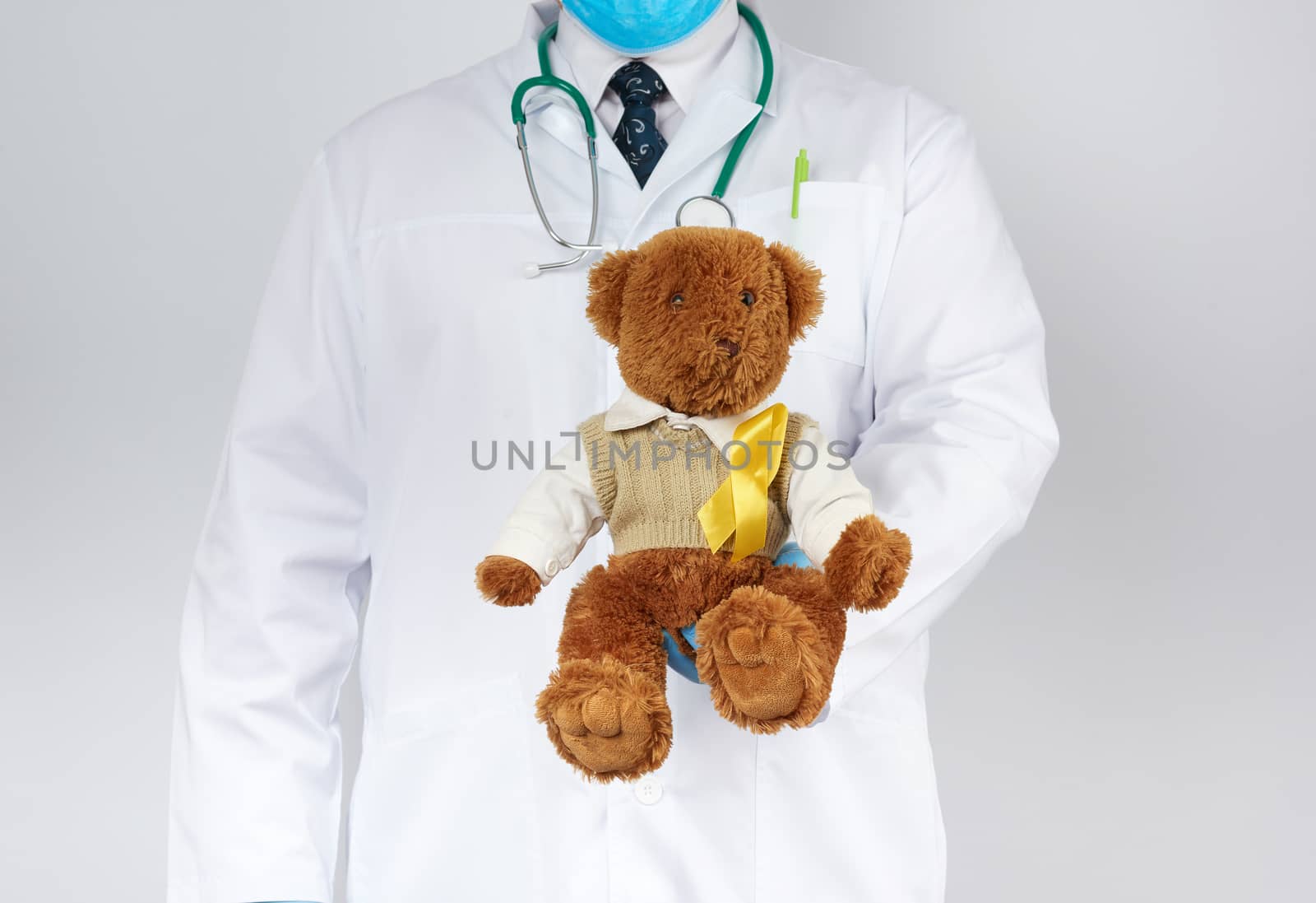 pediatrician in white coat, blue latex gloves holds a brown teddy bear with a yellow ribbon on a sweater, concept of the fight against childhood cancer, problem of suicides