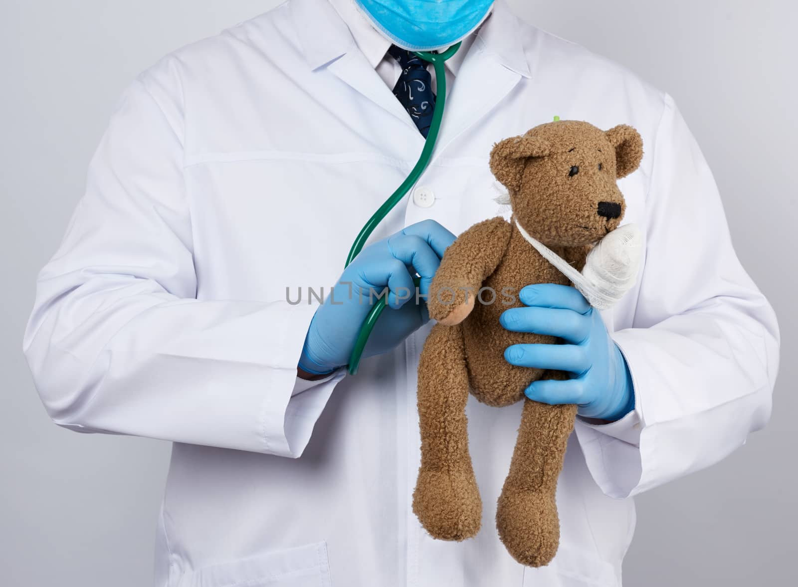 pediatrician in a white coat, blue latex gloves holds a brown teddy bear with a bandaged paw, doctor examines the toy with a stethoscope, white background