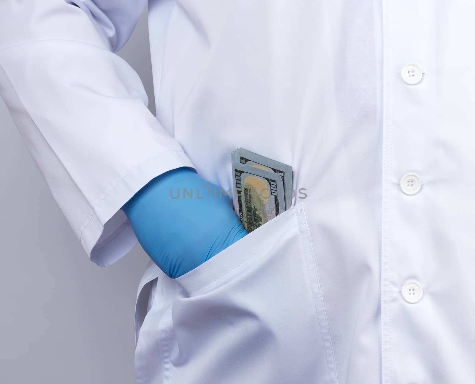 male doctor in a white medical coat and blue latex gloves puts a stack of US dollars in his coat pocket, concept of a bribe, close up