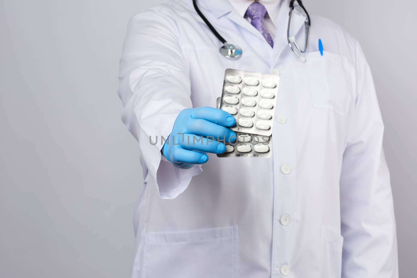adult doctor therapist is dressed in a white uniform coat and blue sterile gloves is standing and holding a stack of pills in blister packs, white background