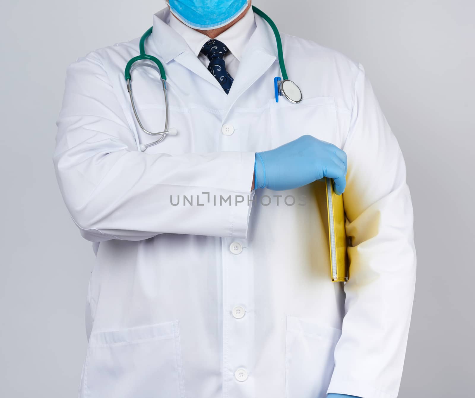 male doctor standing in a white medical coat and blue latex gloves holds a yellow notebook on a white background, therapist
