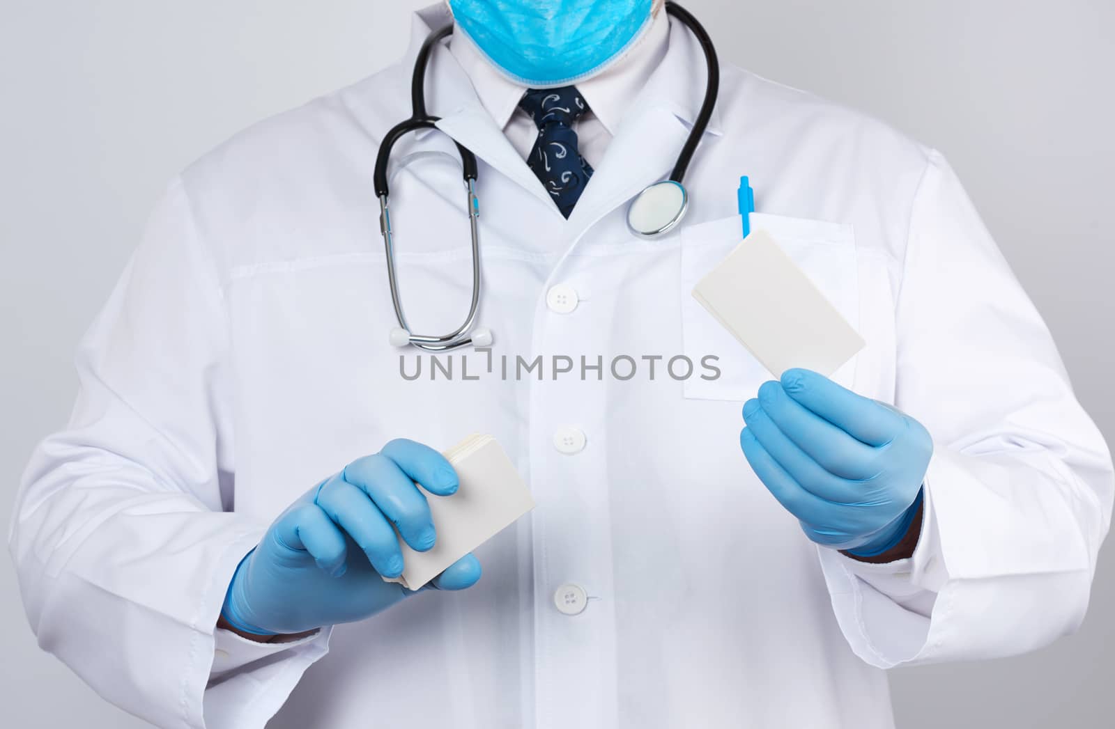adult doctor therapist is dressed in a white robe uniform and blue sterile gloves is standing and holding a stack of empty white paper business cards, place for inscription