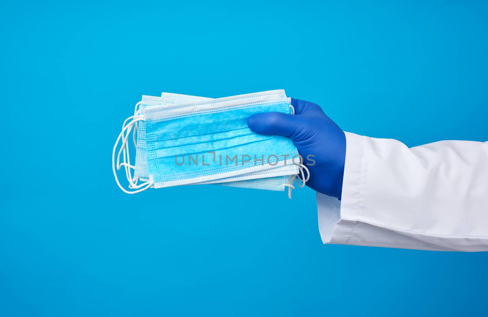 doctor in a white coat, blue latex sterile gloves holds textile disposable medical masks in his hand on a blue background