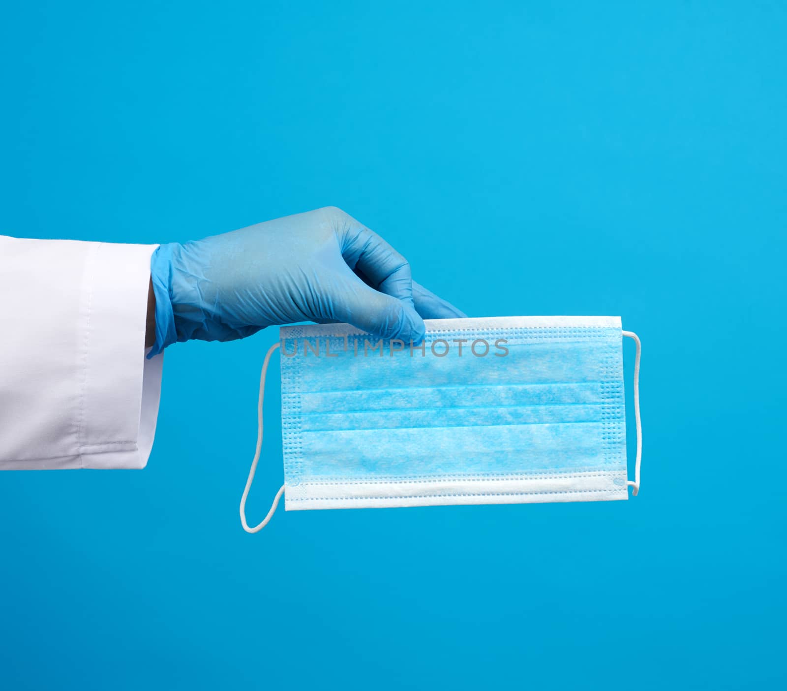 doctor in blue latex sterile gloves holds a blue disposable mask made of non-woven material on a blue background, protective accessory for the respiratory tract from the virus