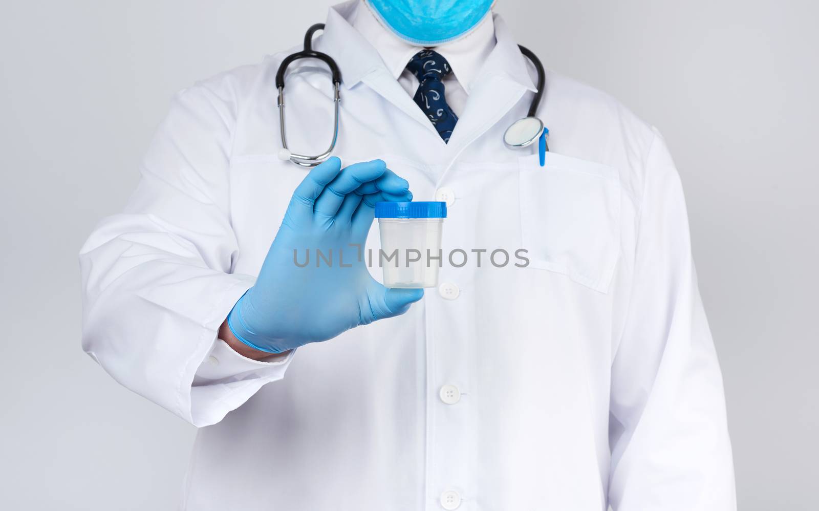 male doctor in a white coat and tie stands and holds a plastic container for urine specimen on a white background, wearing blue sterile medical gloves