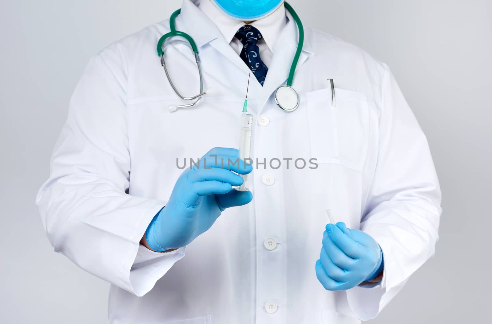 male doctor in a white coat and tie stands and holds a plastic syringe with a needle on a white background, wearing blue sterile medical gloves in his hands the concept of vaccination against diseases