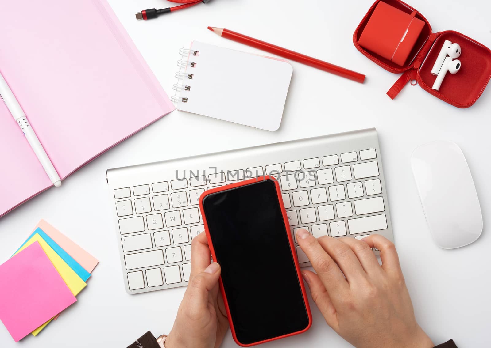 hands holding a smartphone with a blank black screen over a white desktop with a keyboard, computer mouse, notebook and color stickers, top view, workplace
