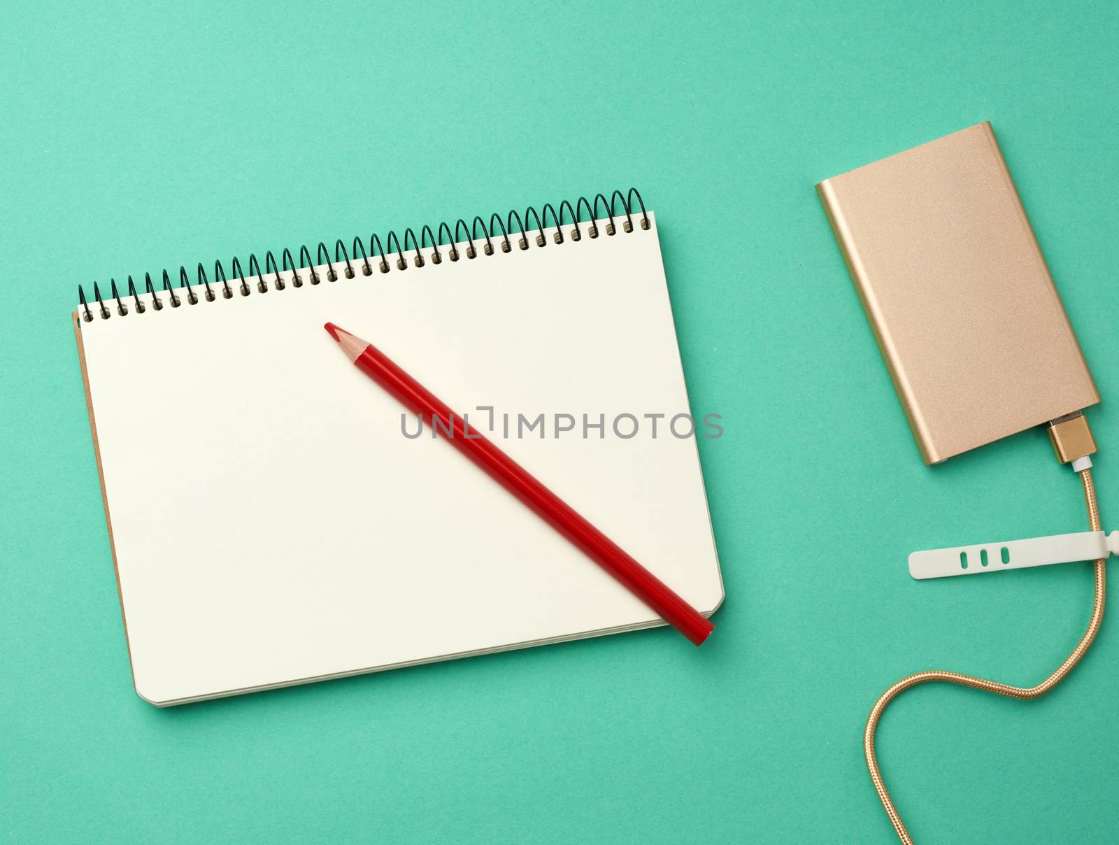 open notebook with blank white sheets and golden power bank and cord with a usb connector for recharging mobile devices on a green background, top view