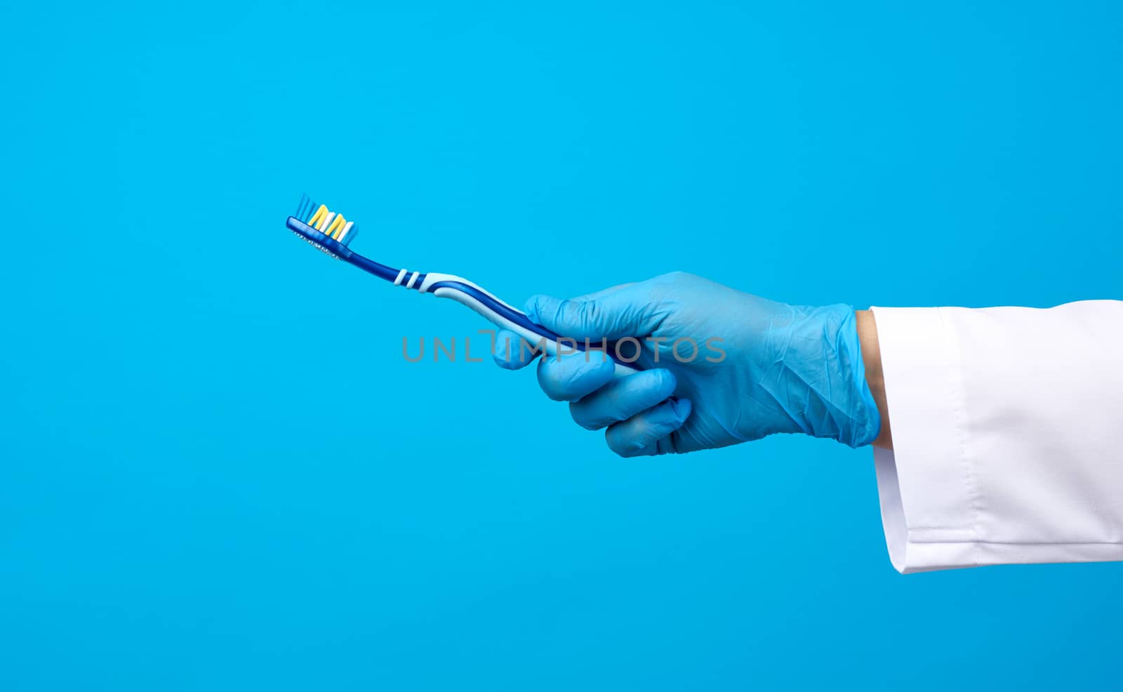 doctor therapist is dressed in a white robe uniform and blue sterile gloves holding a toothbrush, daily brushing concept, blue background