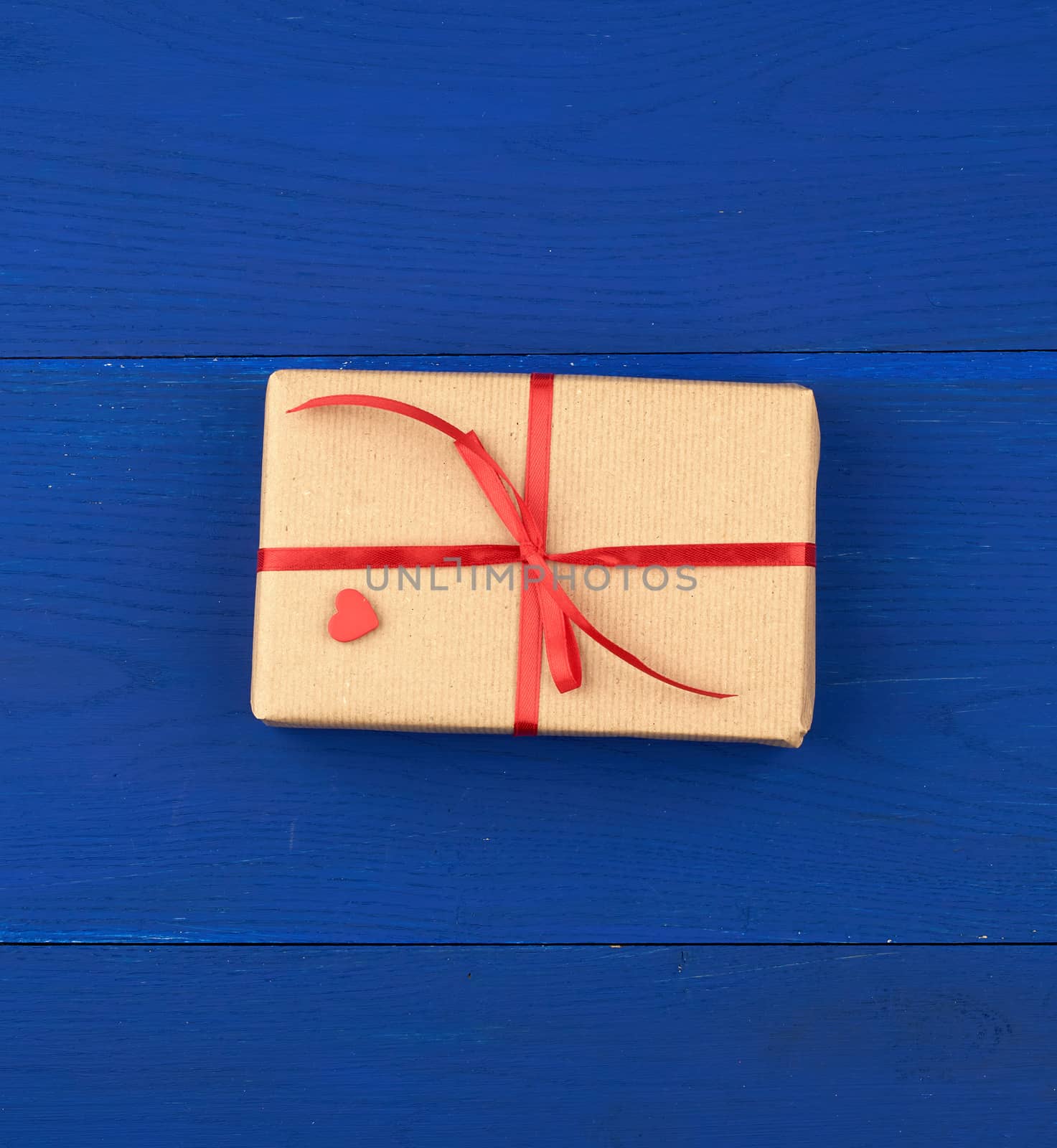 gift wrapped in brown kraft paper and tied with a thin red ribbon on a blue wooden background, top view, flat lay. Eco package design