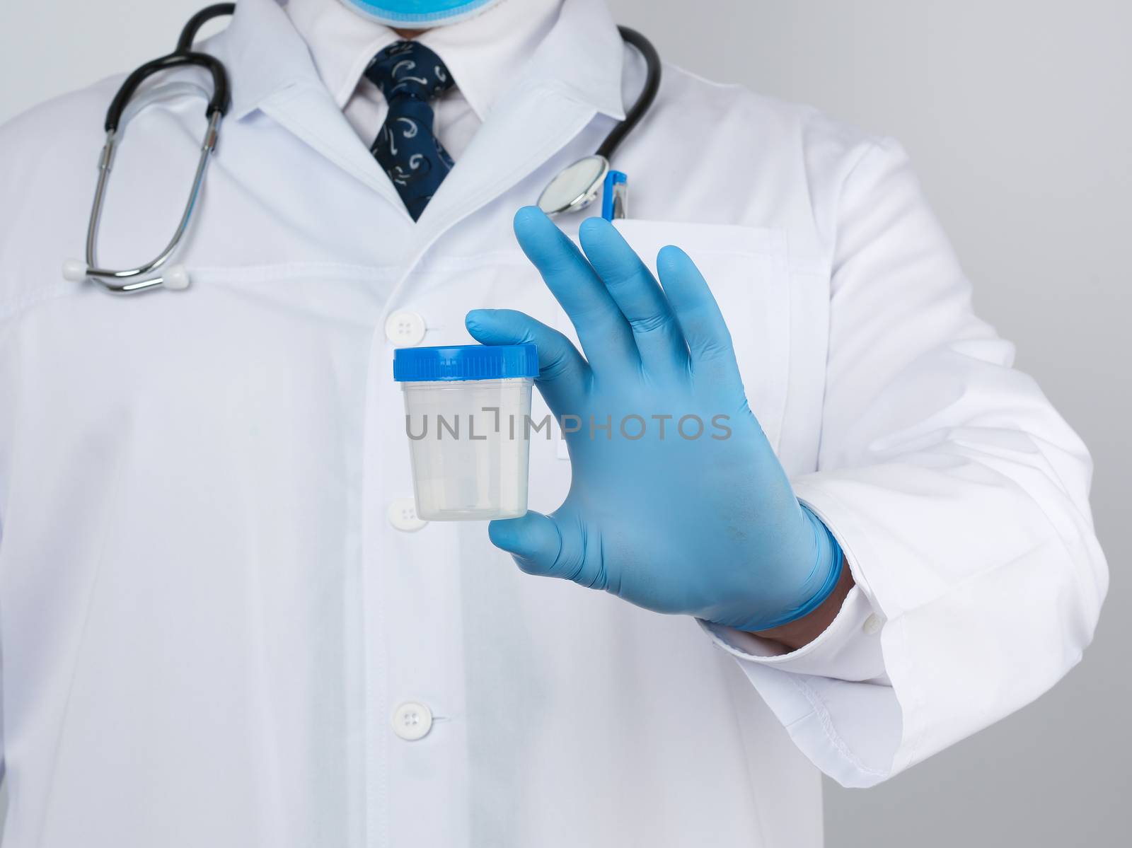 male doctor in a white coat and tie stands and holds a plastic container for urine specimen, wearing blue sterile medical gloves