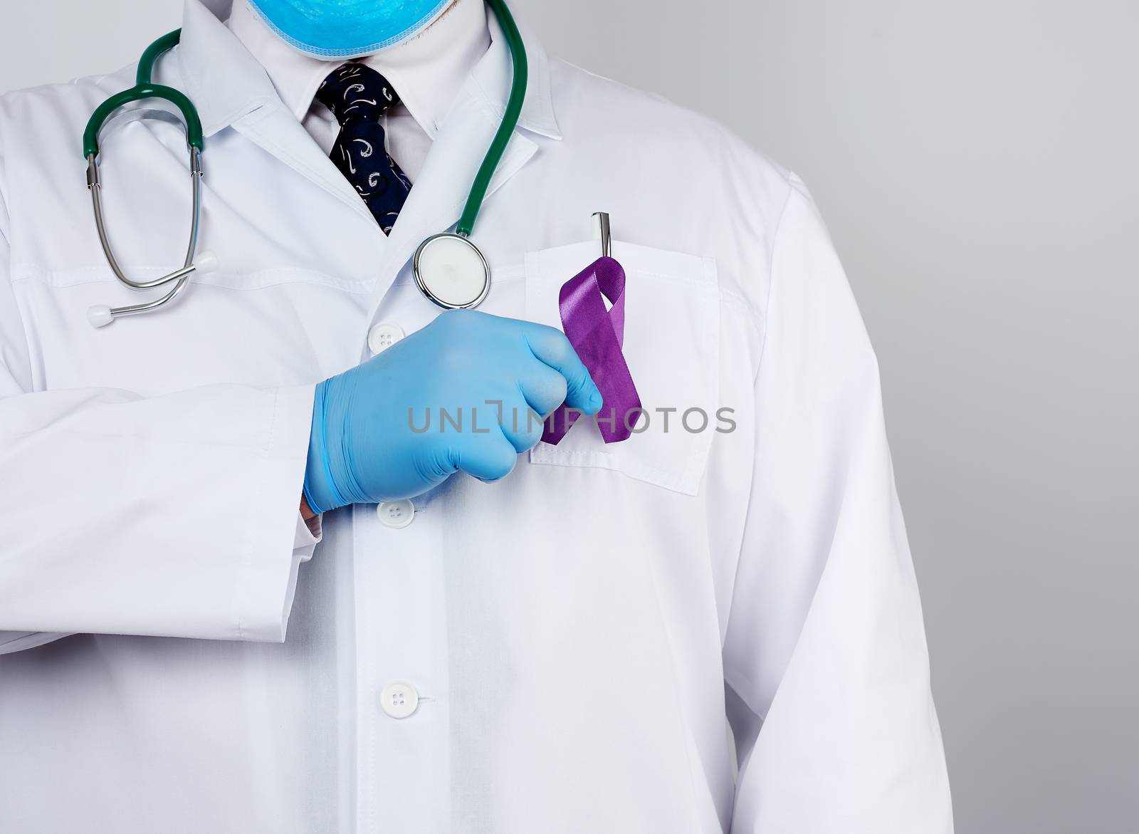 male doctor in a white coat and tie stands and holds a purple silk ribbon in the form of a loop, wearing blue sterile medical gloves. Symbol of early research and disease control, Alzheimer's disease, cystic fibrosis, lupus, pancreatic cancer, sarcoidosis