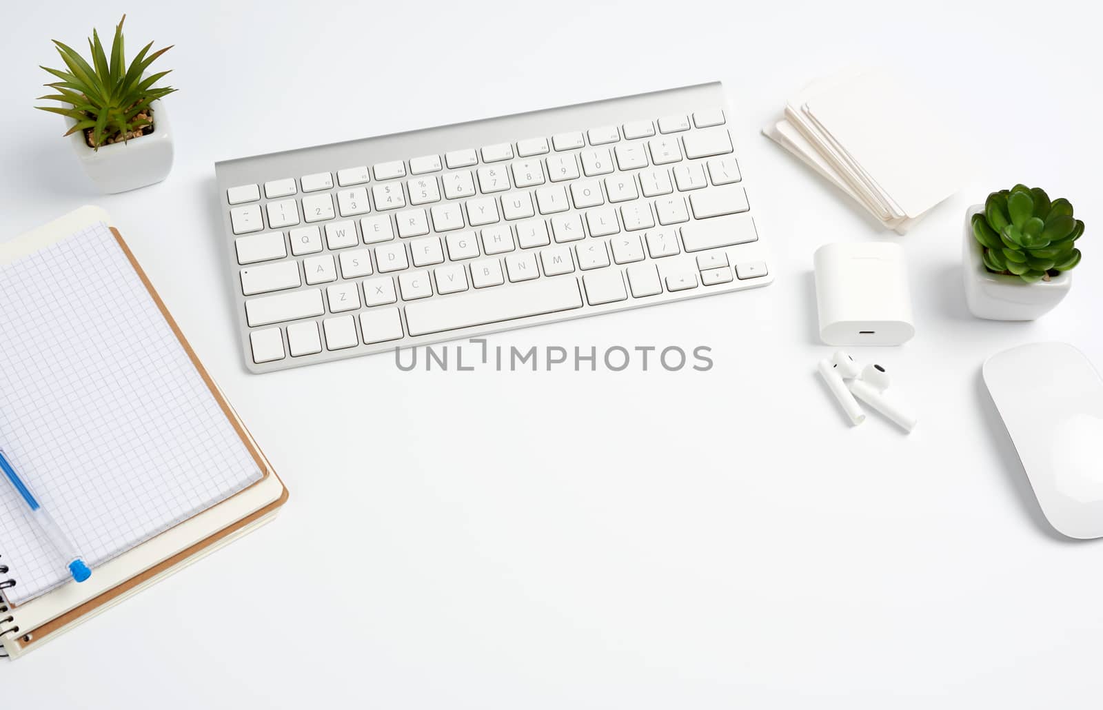 White wireless keyboard, a stack of notebooks, green plants in pots and a mouse, workplace of a freelancer, businessman. White table