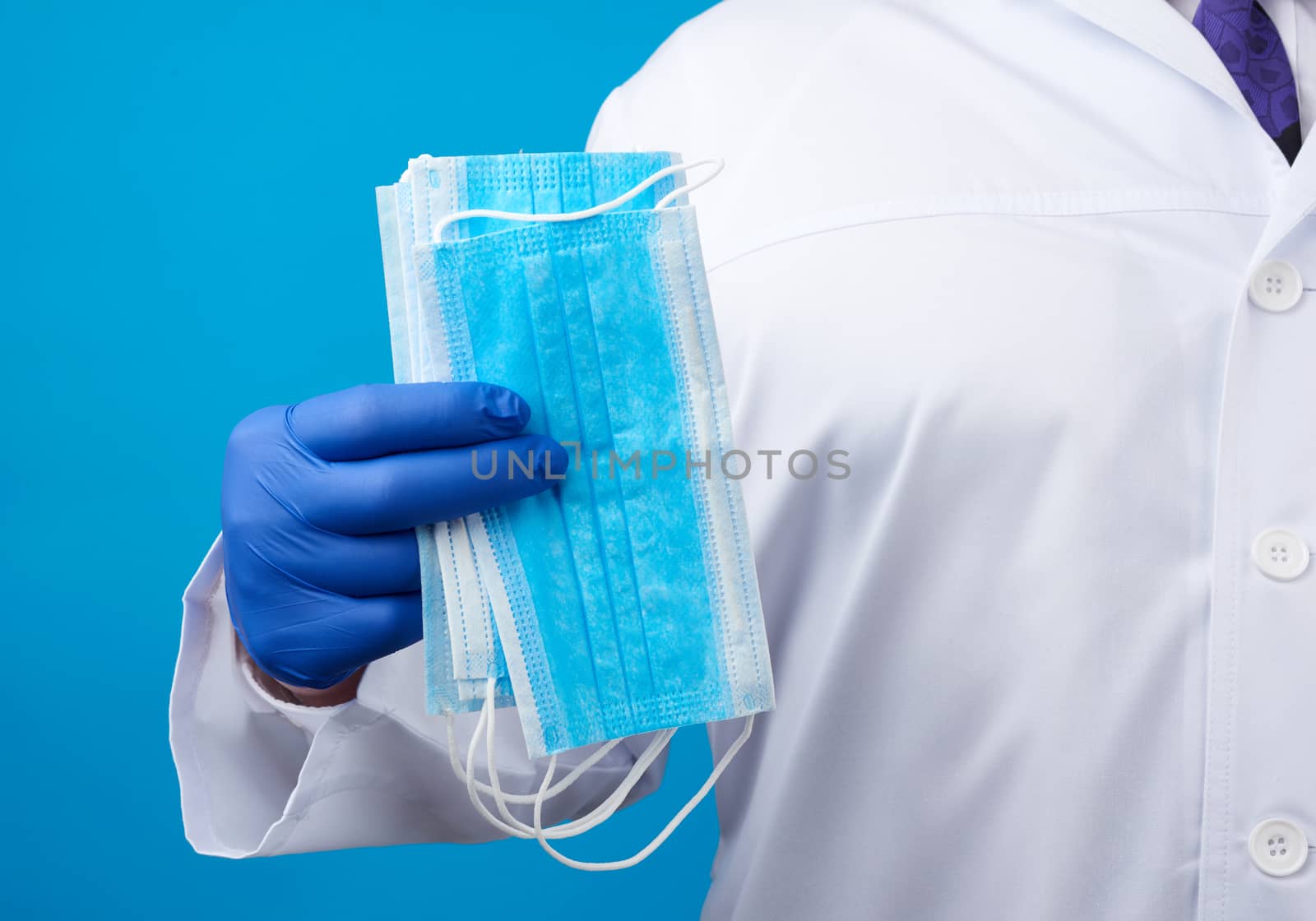 doctor in a white coat, blue latex sterile gloves holds textile disposable medical masks in his hand on a blue background