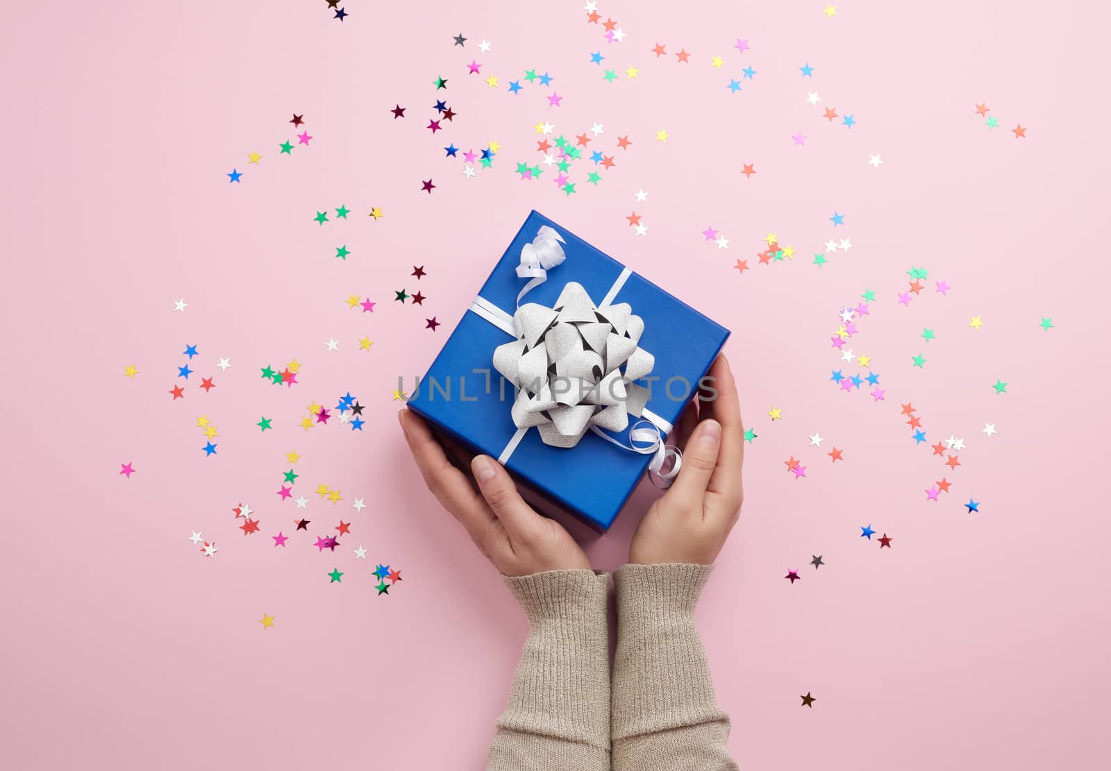 two female hands in a sweater hold closed square blue box with a bow on a pink background, top view, festive backdrop