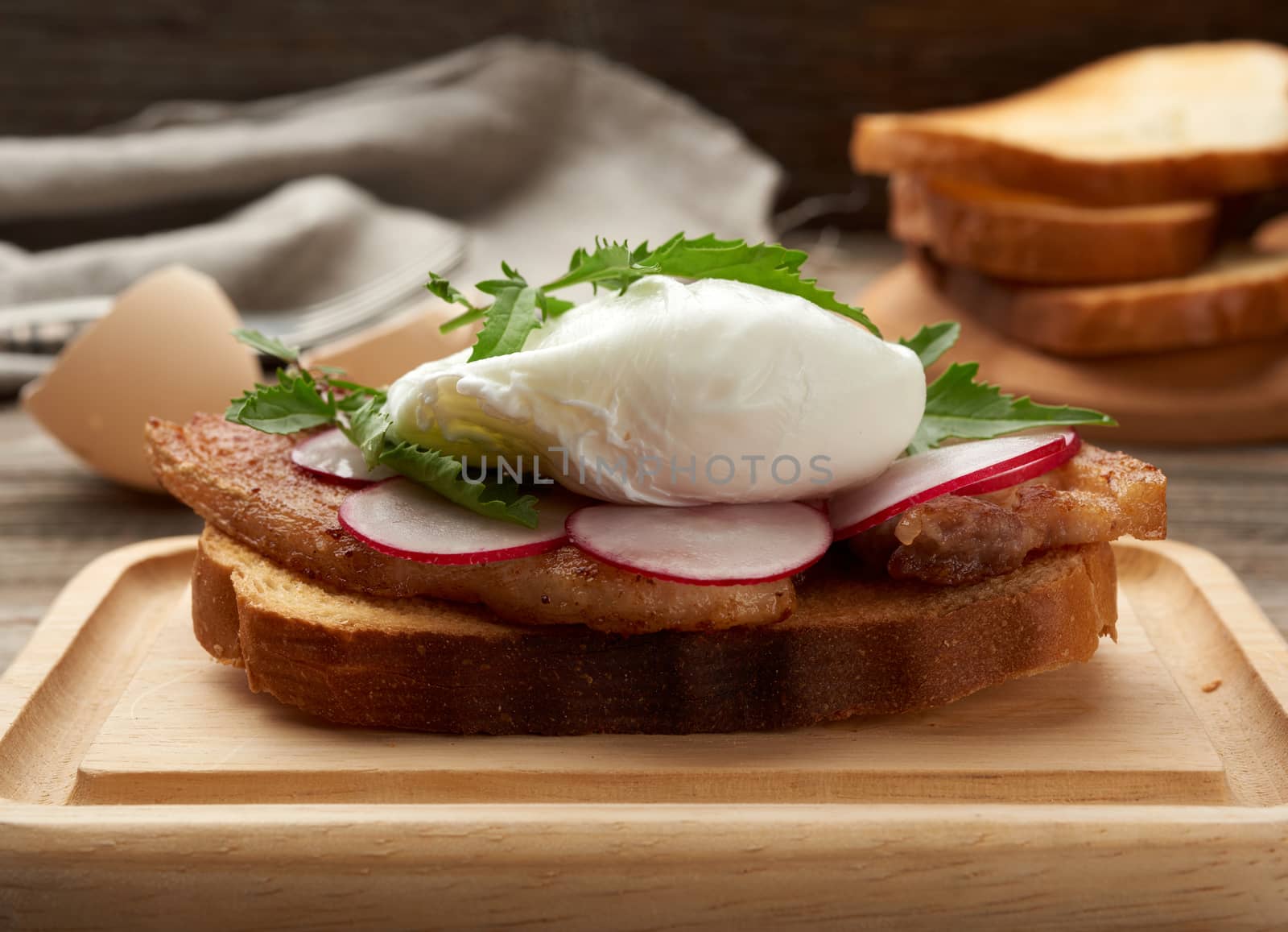 sandwich on toasted white slice of bread with poached eggs, green leaves of arugula and radish, morning breakfast on a brown wooden board