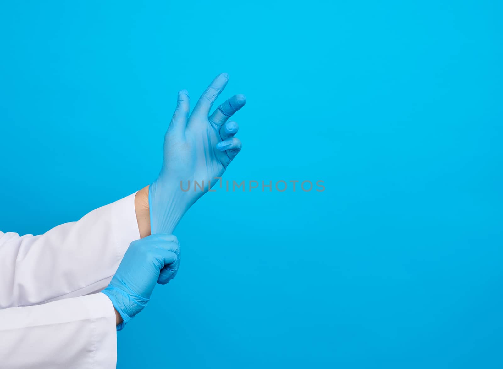 female doctor in a white coat puts on her hands blue latex sterile medical gloves on her hands, blue background, place for text