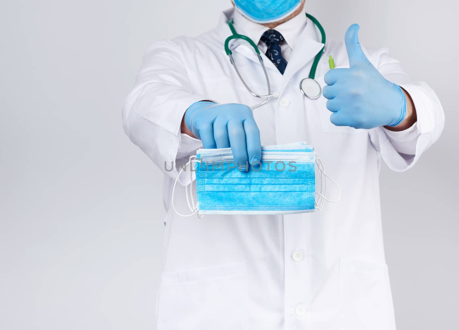 doctor in a white coat, blue latex sterile gloves holds textile medical masks in his hand on a white background, other hand shows like, protective accessory against viruses and bacteria
