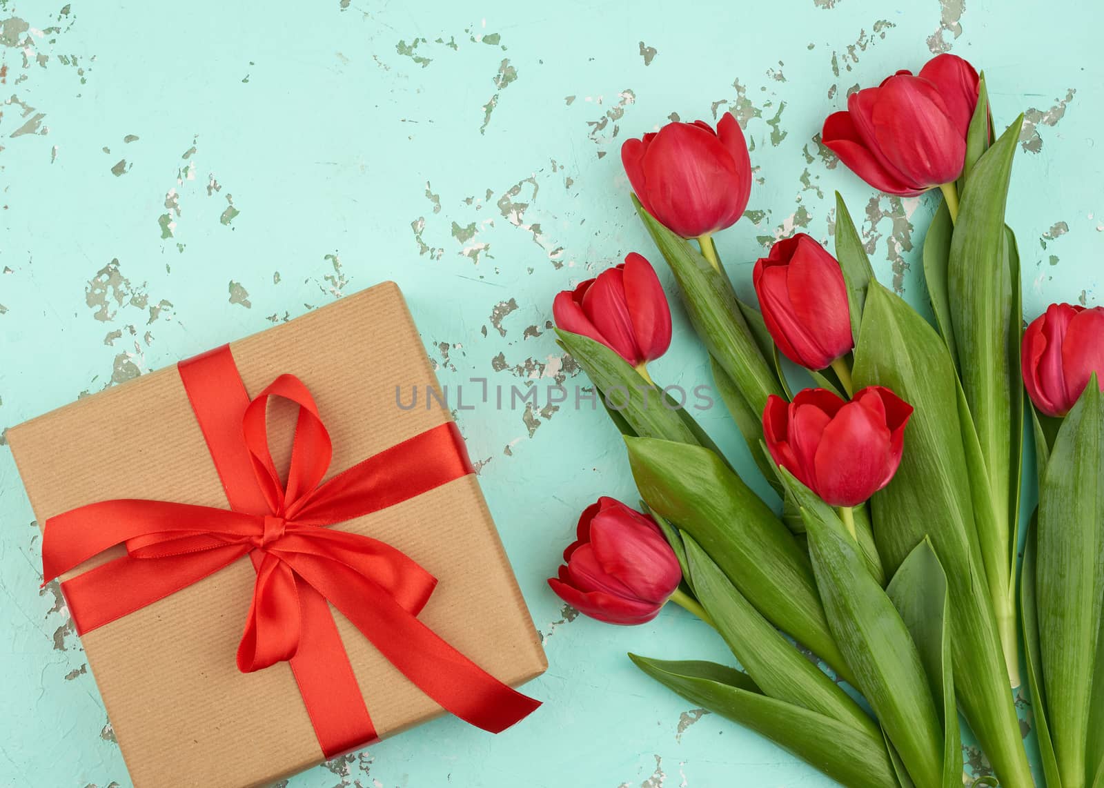 bouquet of red blooming tulips with green leaves, wrapped gift in brown craft paper and tied with a silk red ribbon on a green background, top view. Festive backdrop for birthday, Valentine's day