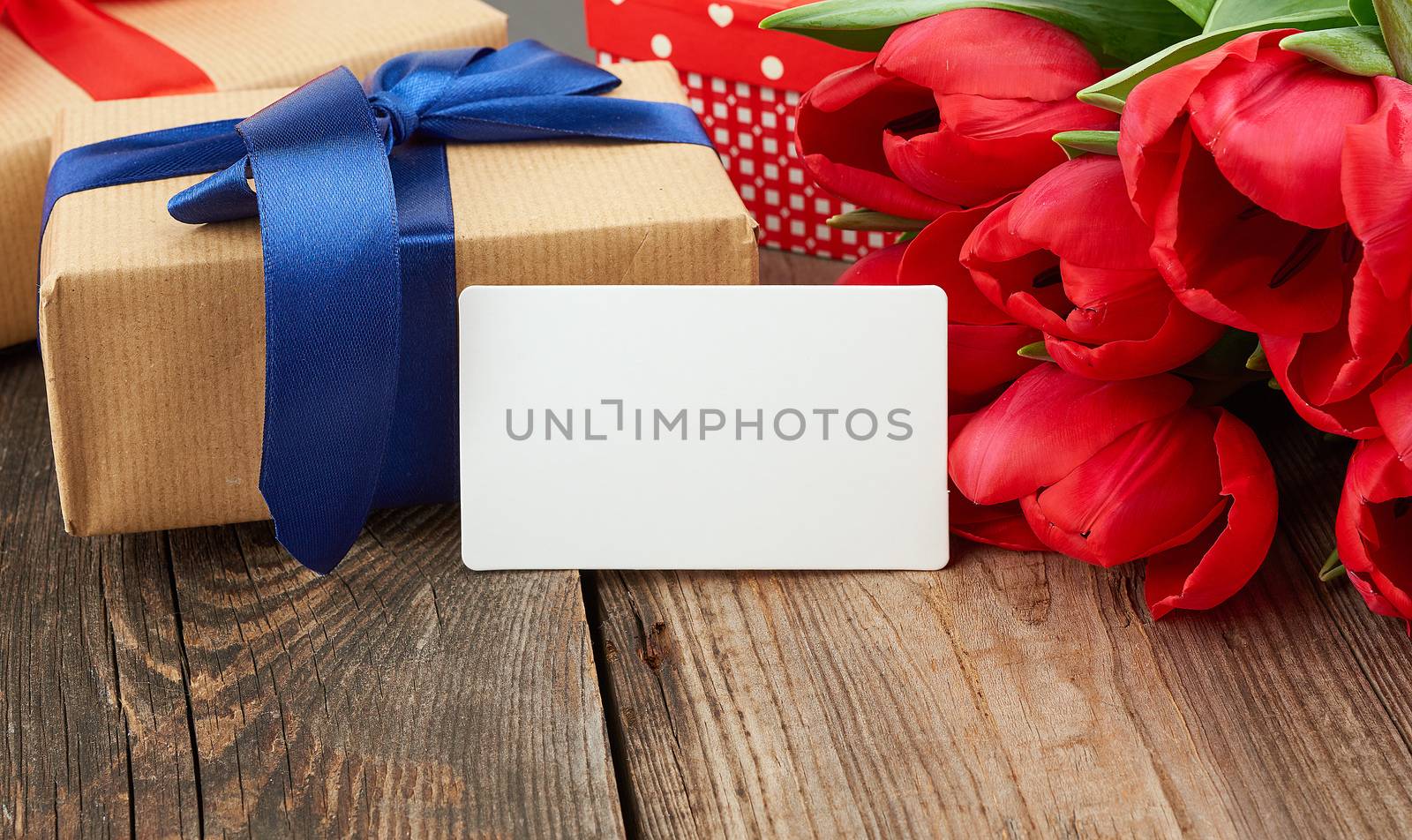 empty rectangular white paper business card, a bouquet of red tulips and wrapped gifts on a gray wooden table. Festive backdrop for birthday, Valentine's day, anniversary