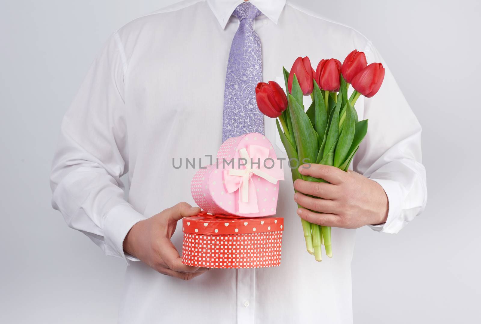 adult man in a white shirt and a lilac tie holding a bouquet of red tulips with green leaves and gift box on a white background, concept for happy birthday, anniversary, valentines day, date