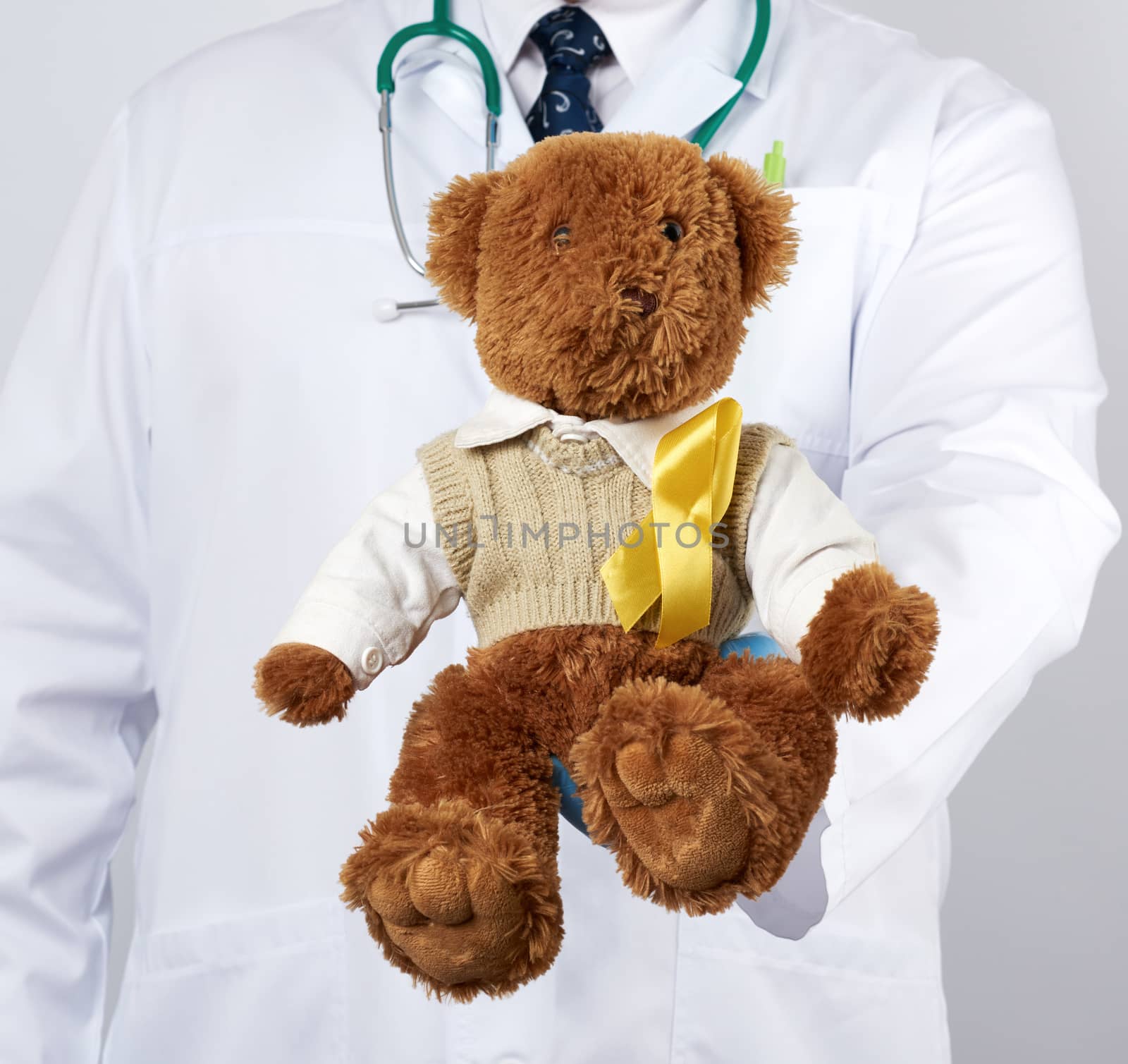 pediatrician in white coat, blue latex gloves holds a brown teddy bear with a yellow ribbon on a sweater, concept of the fight against childhood cancer, problem of suicides