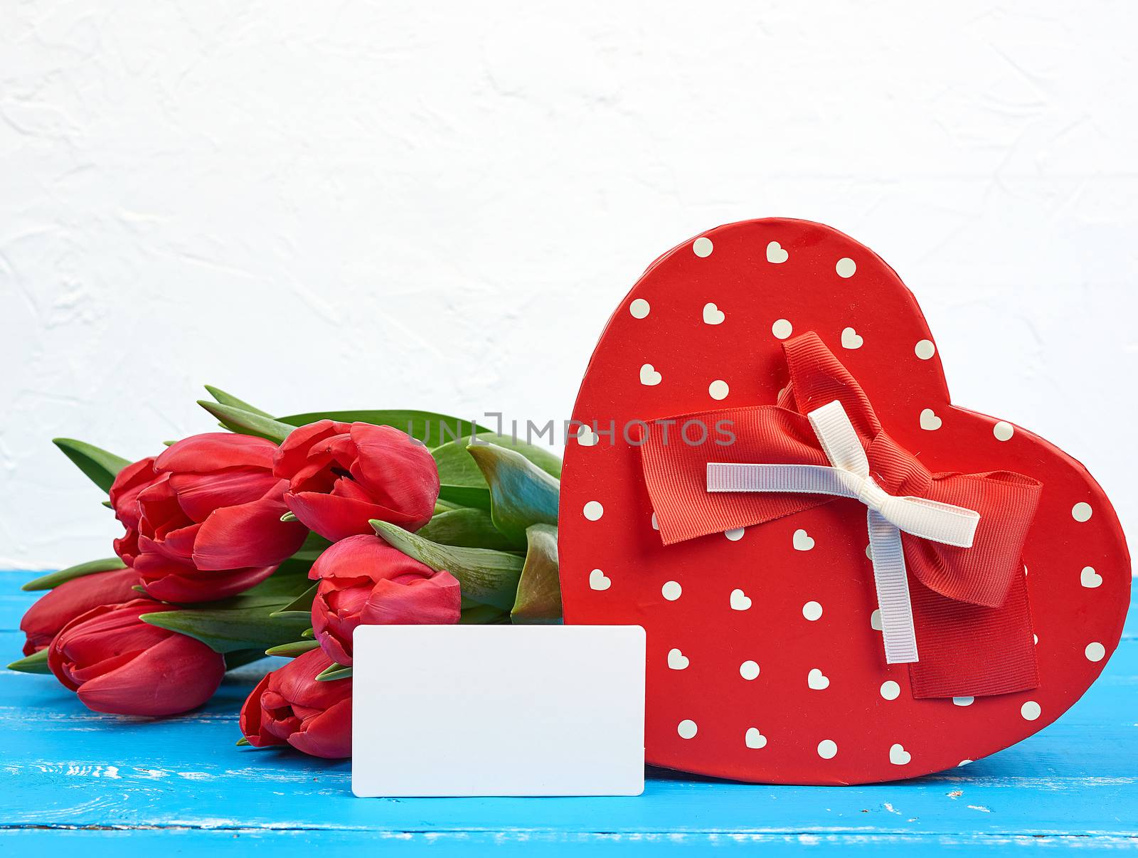 empty rectangular white paper business card, bouquet of red tulips and a heart-shaped cardboard box on a blue wooden background. Festive backdrop for birthday, Valentine's day, anniversary