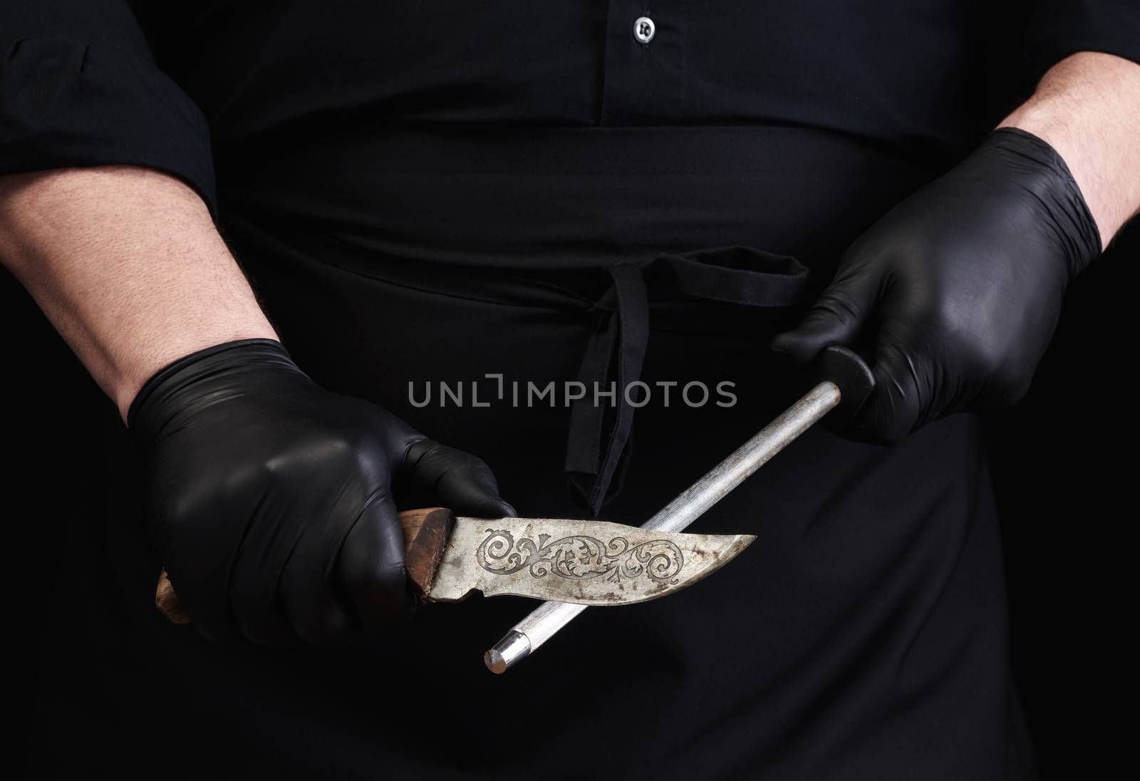 chef in a black shirt and black latex gloves sharpen a kitchen knife on an iron sharpener with a handle, low key