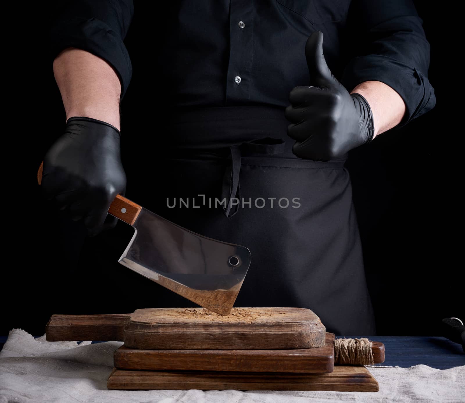 male cook in black uniform and black latex gloves holds a large sharp meat knife over a cutting board, another hand shows like gesture, black background