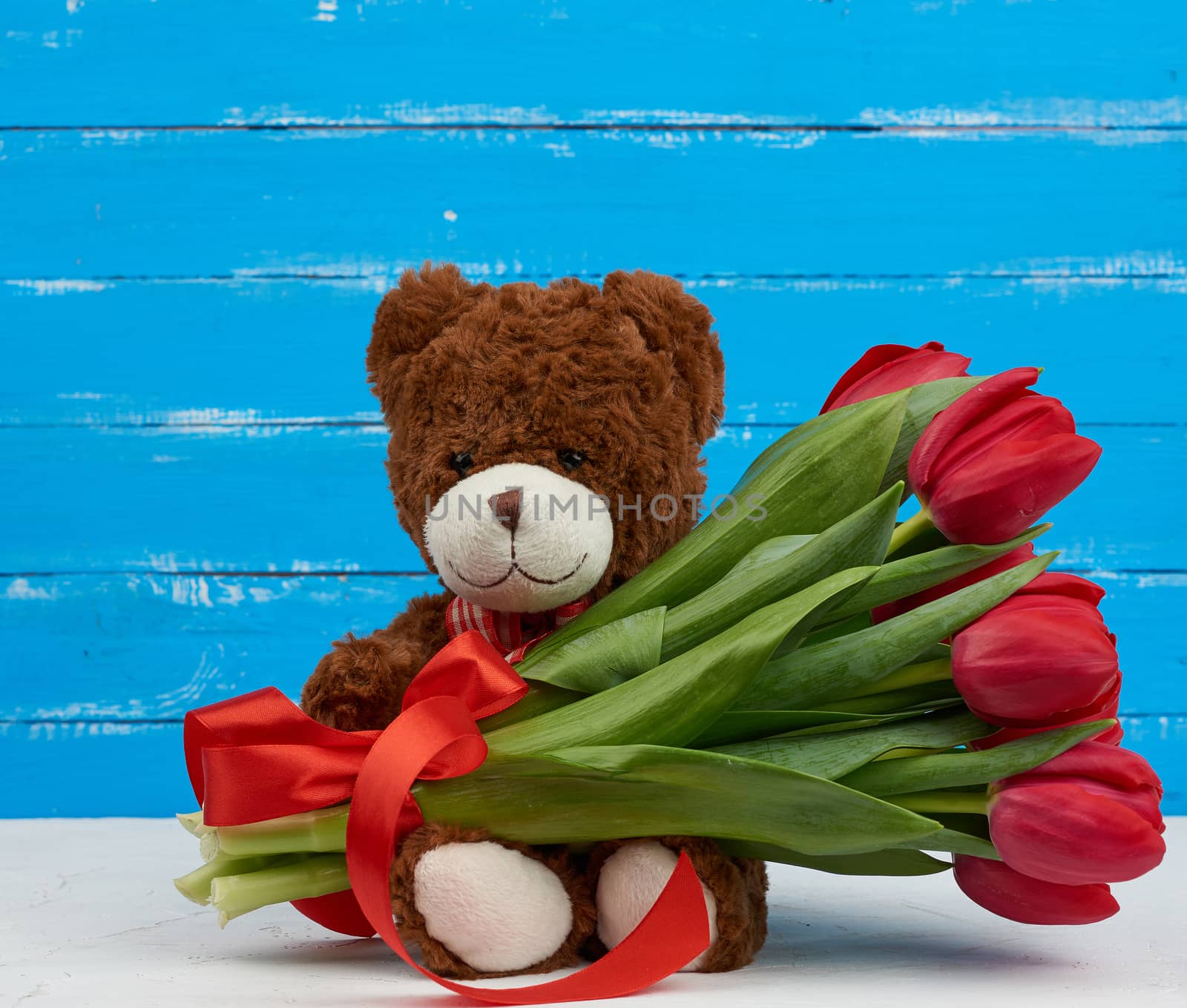 cute brown teddy bear with patches sits on a blue wooden background and holds in his paw a bouquet of red tulips, festive backdrop for birthday, Valentine's day, anniversary